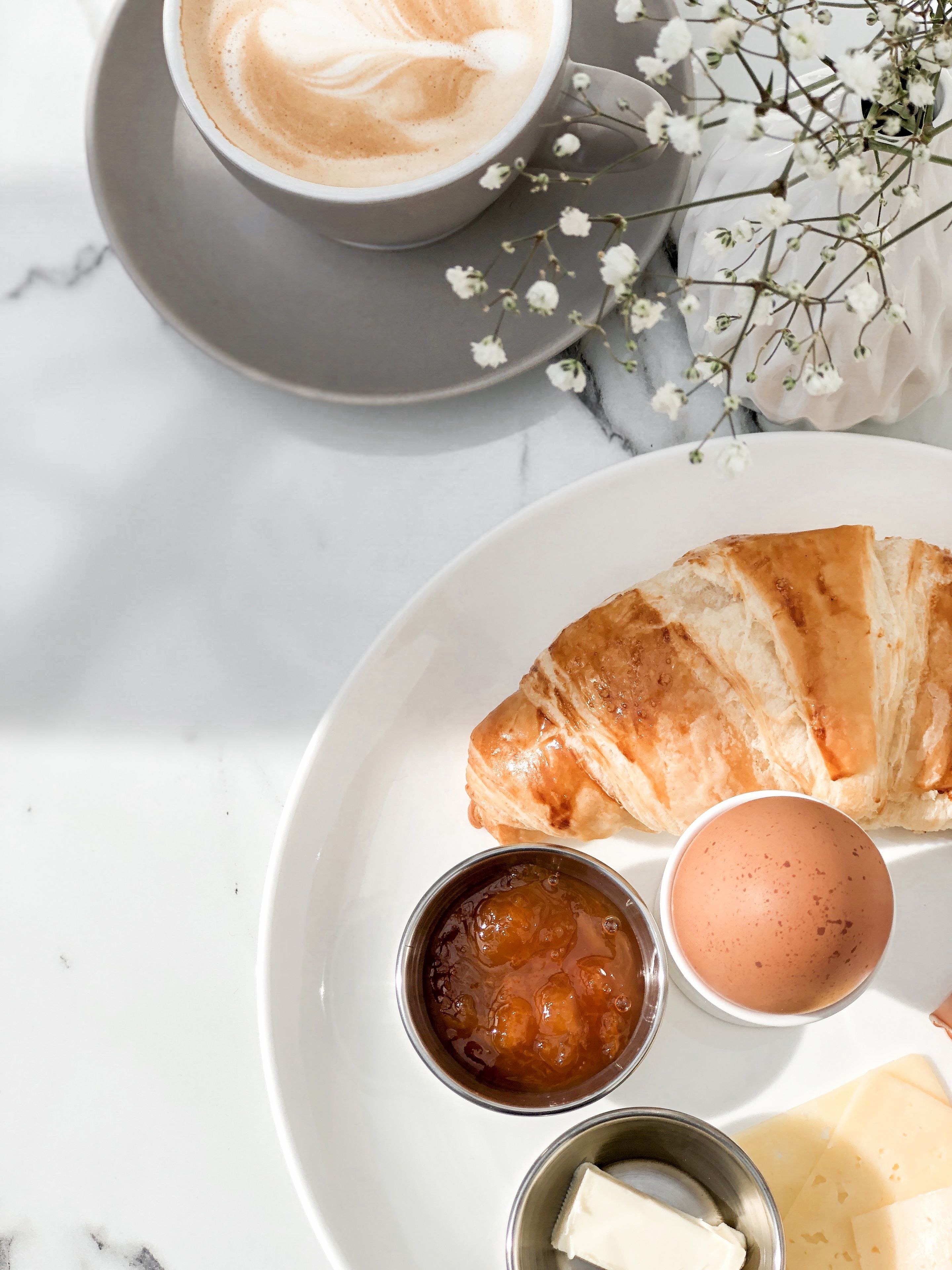 Een ontbijttafel met een croissant, een kop koffie, een klein schaaltje jam, een klein schaaltje boter en een gekookt ei op een wit bord.