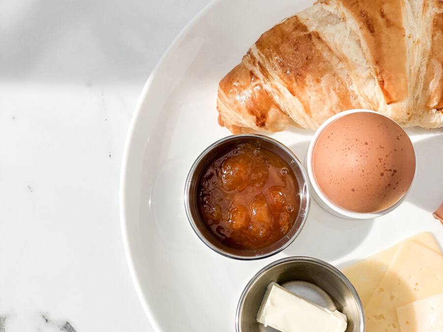 A breakfast setting with a croissant, a cup of coffee, a small bowl of jam, a small bowl of butter, and a boiled egg on a white plate.