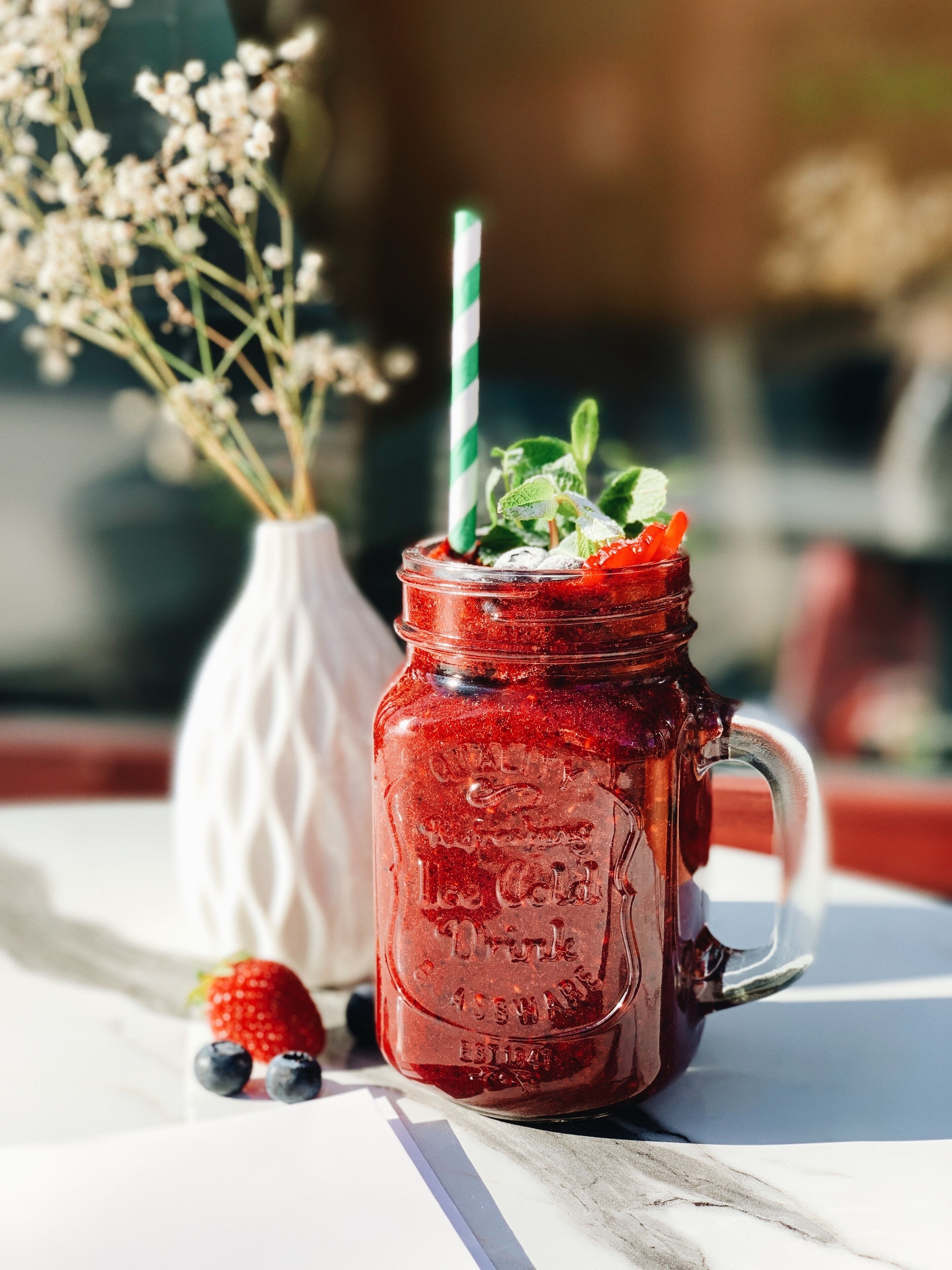 Un bocal en verre avec une paille à rayures vertes et blanches, des feuilles de menthe, des fraises et des myrtilles, le tout posé sur une table blanche.