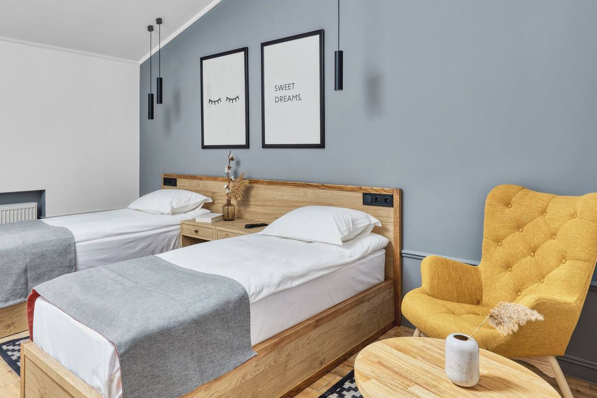 Ein Hotelzimmer mit zwei Einzelbetten, einem Holzkopfende, einem gelben Sessel und einem kleinen Tisch mit einer Vase.