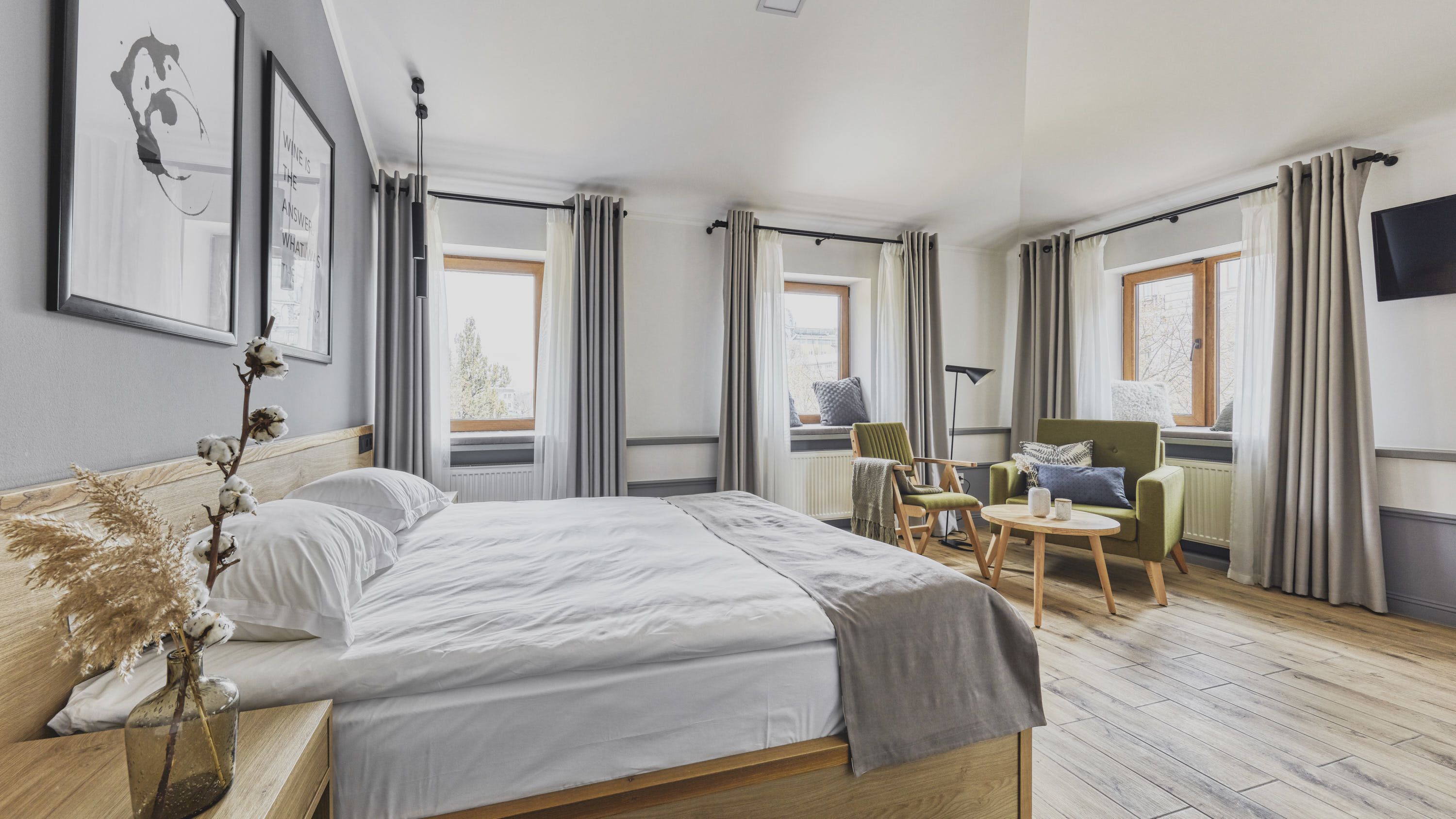 Una habitación de hotel moderna con una cama grande, un área de descanso con una silla y una mesita, y ventanas amplias con cortinas.