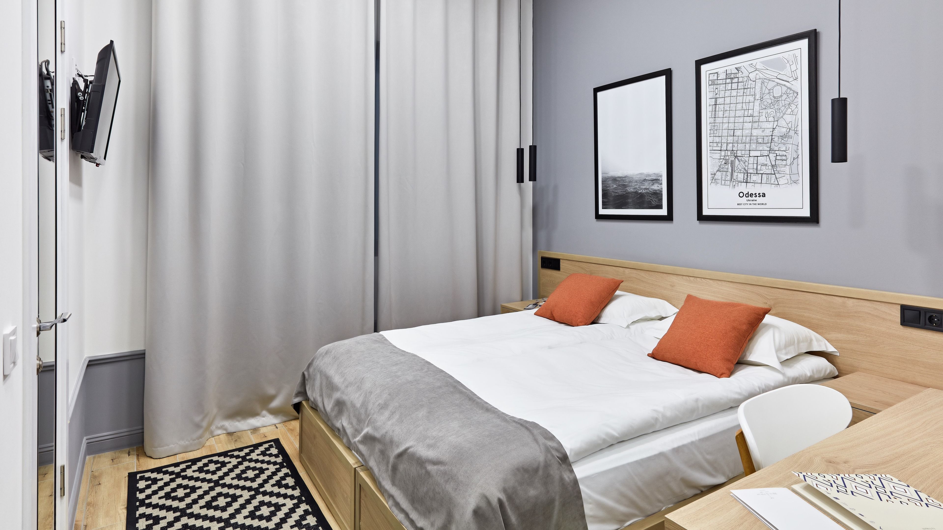 A neatly arranged hotel room with a bed, a desk, and a chair. There are curtains, a framed map, and a wall-mounted light fixture.