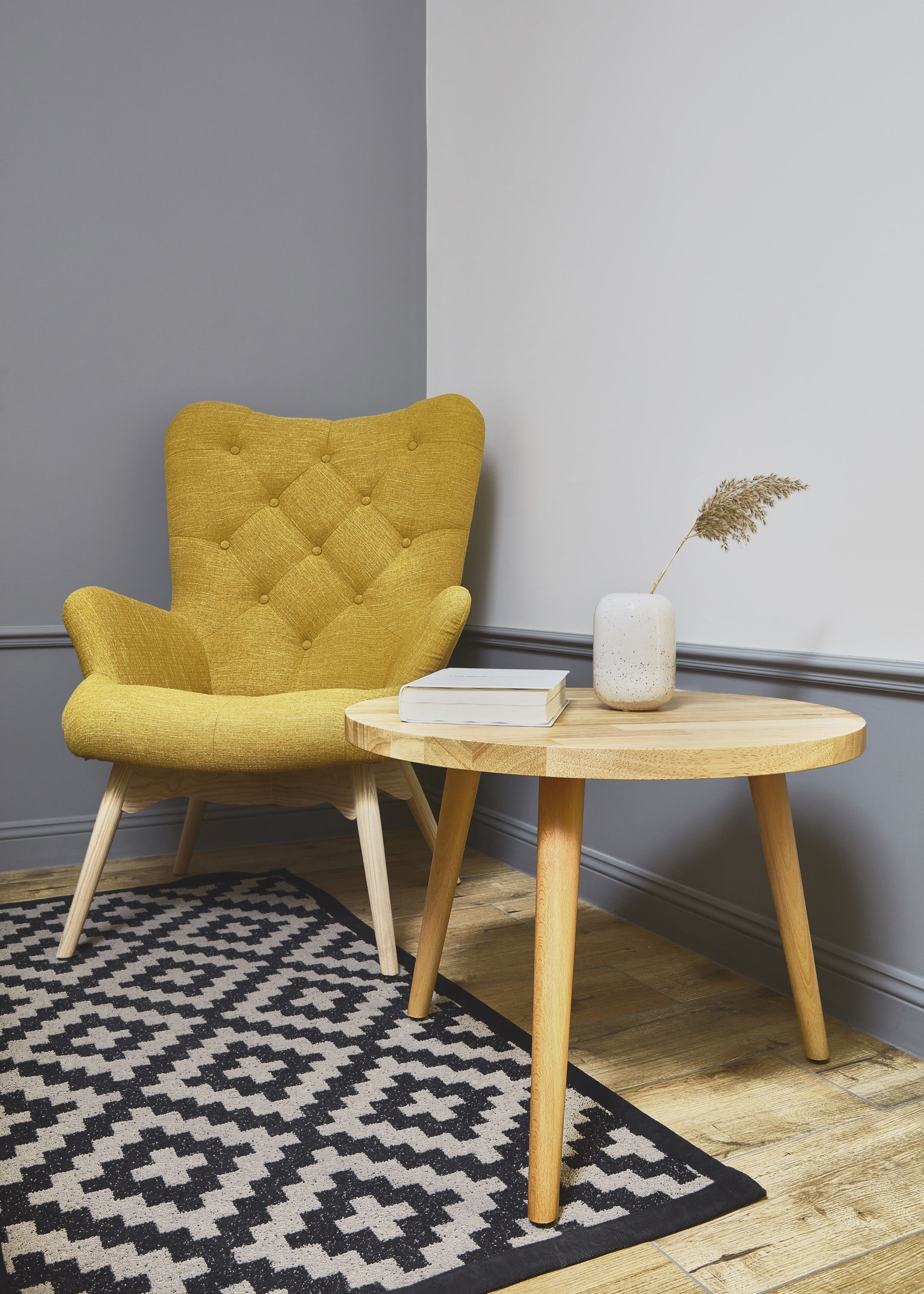 A yellow armchair, a round wooden table with a book and a vase with dried plants.