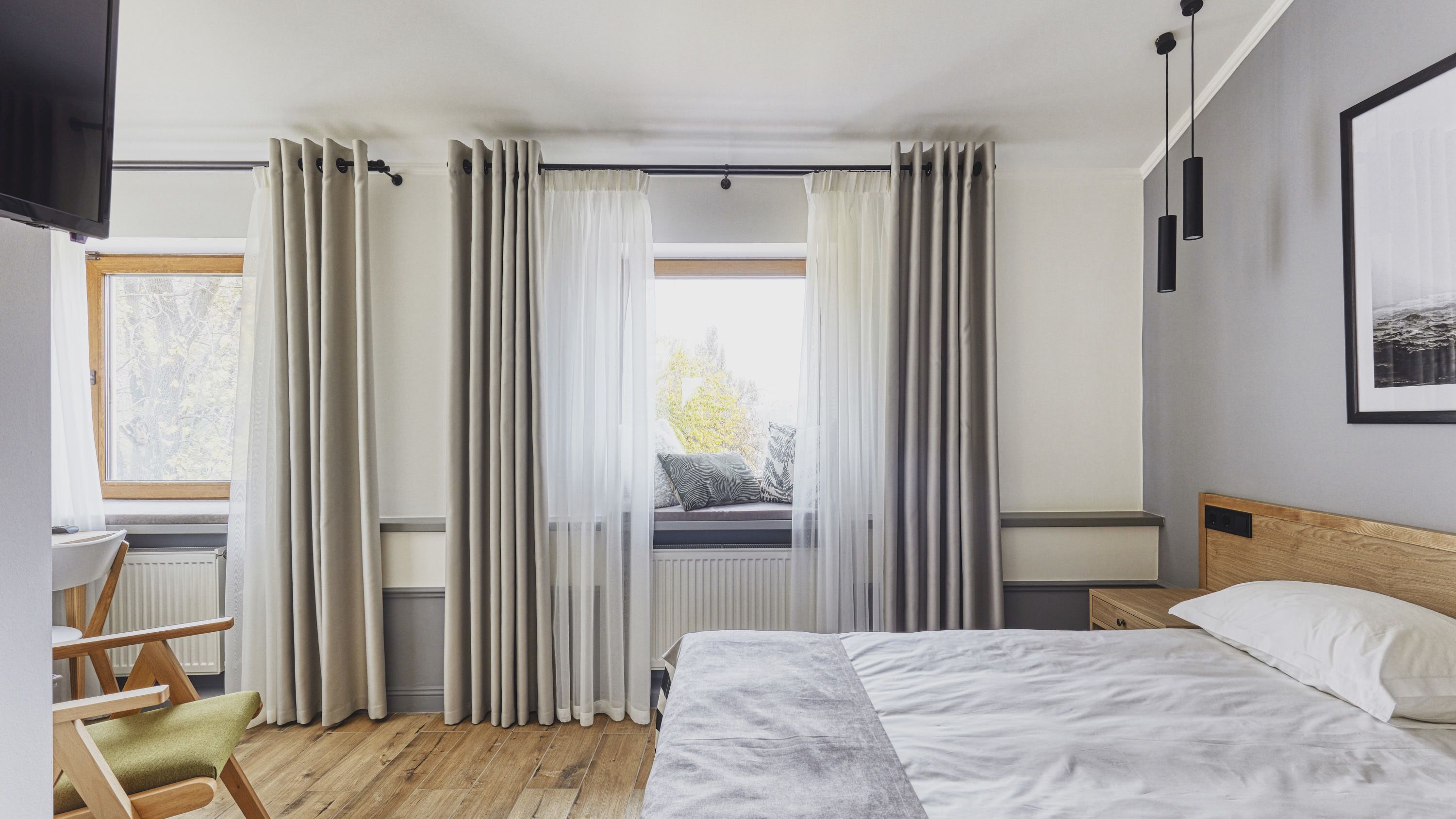 A bedroom with a bed, a window with curtains, a radiator, a wooden headboard, and a chair.