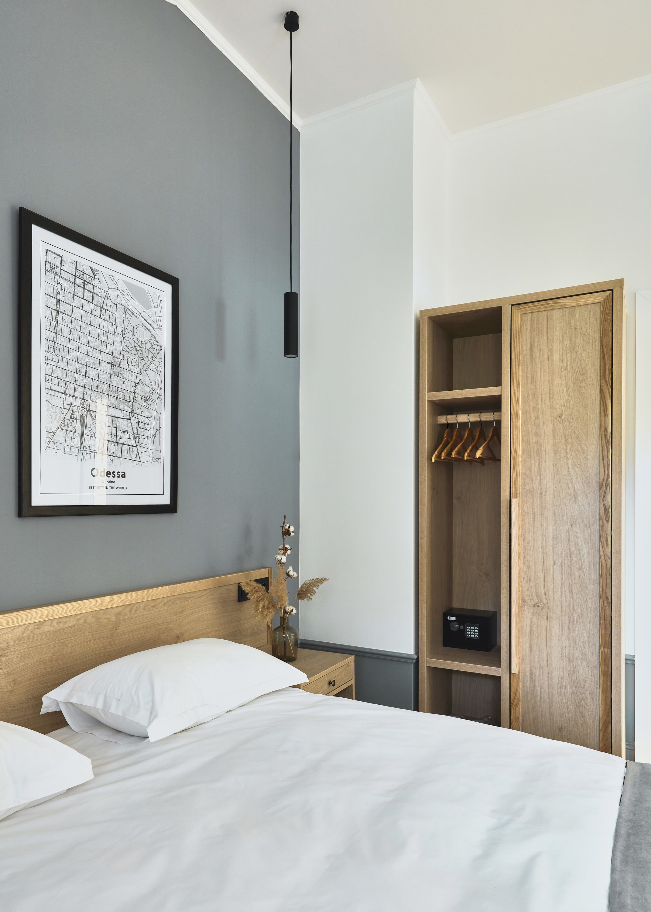 A bedroom with a bed, a wooden headboard, a nightstand, a framed map, a hanging light, and a wardrobe with clothes.