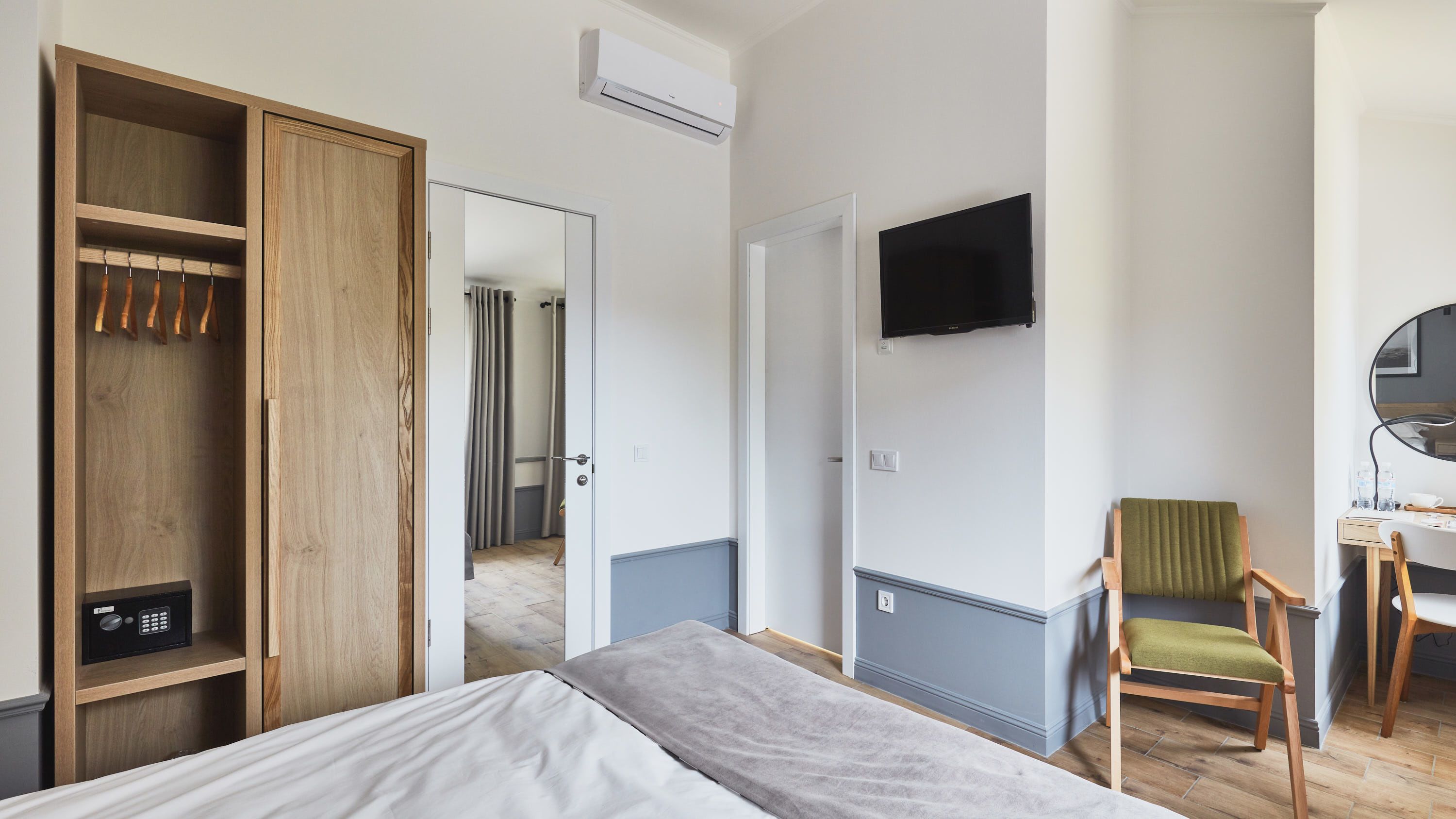 Quarto de hotel com cama, guarda-roupa de madeira, escrivaninha com cadeira, televisão e banheiro visível ao fundo.