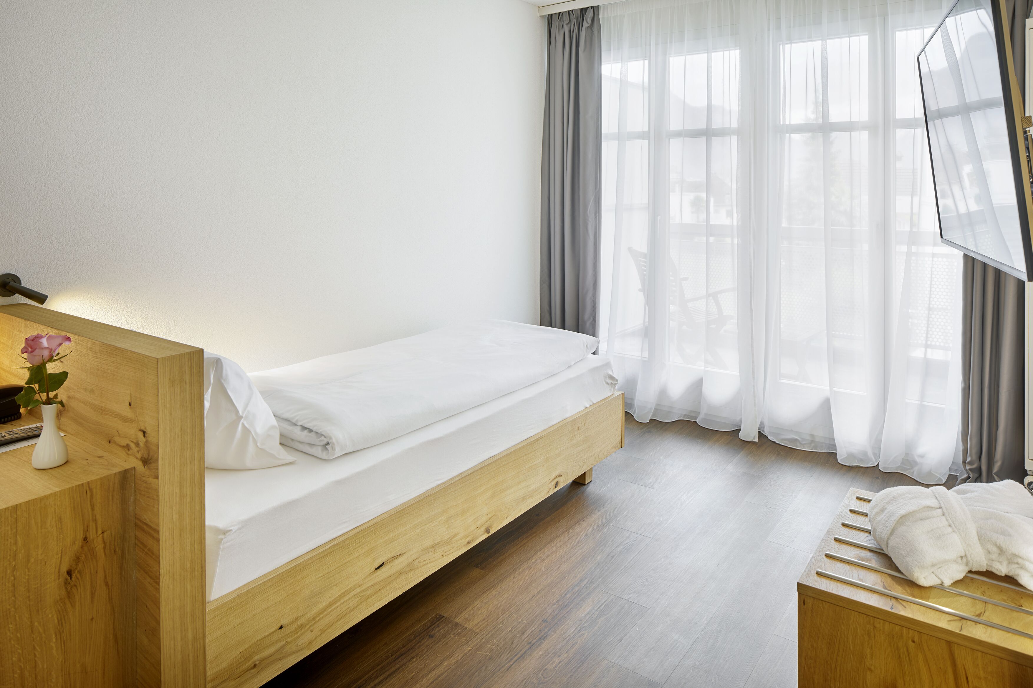Una cama individual con estructura de madera, una mesita de noche con un florero y una ventana con cortinas traslúcidas.