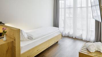 Una cama individual con estructura de madera, una mesita de noche con un florero y una ventana con cortinas traslúcidas.