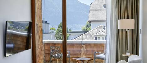 Habitación con puerta de vidrio corrediza que da a un balcón con vistas a montañas y casas. Hay un televisor montado en la pared, una mesa con dos sillas y una lámpara a la derecha.