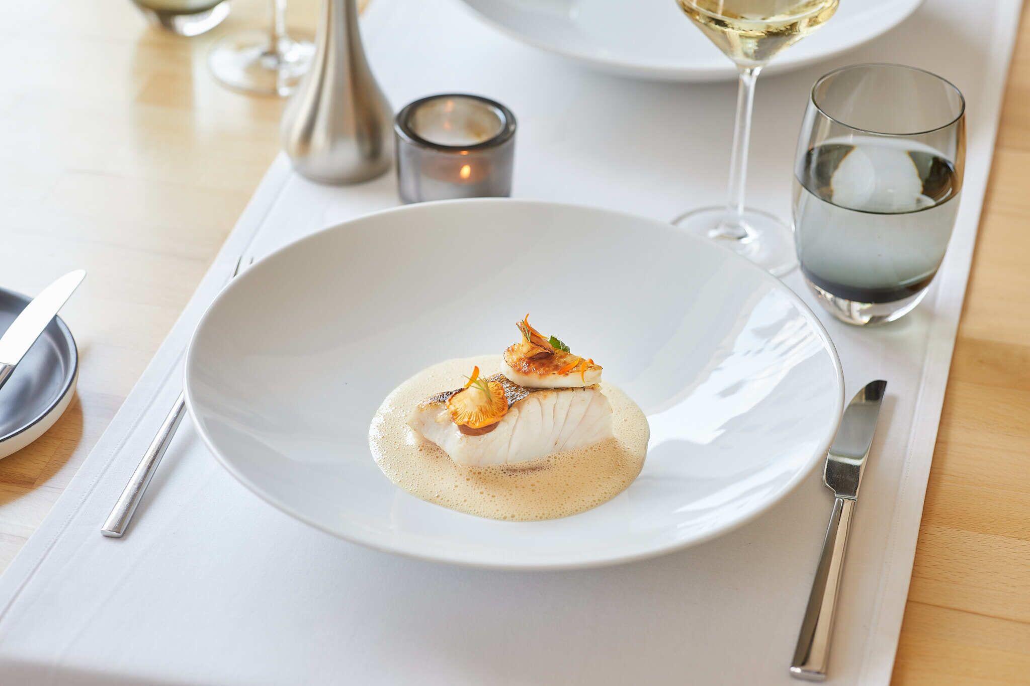 Un plato blanco con mariscos, una copa de vino blanco y una vela sobre una mesa.