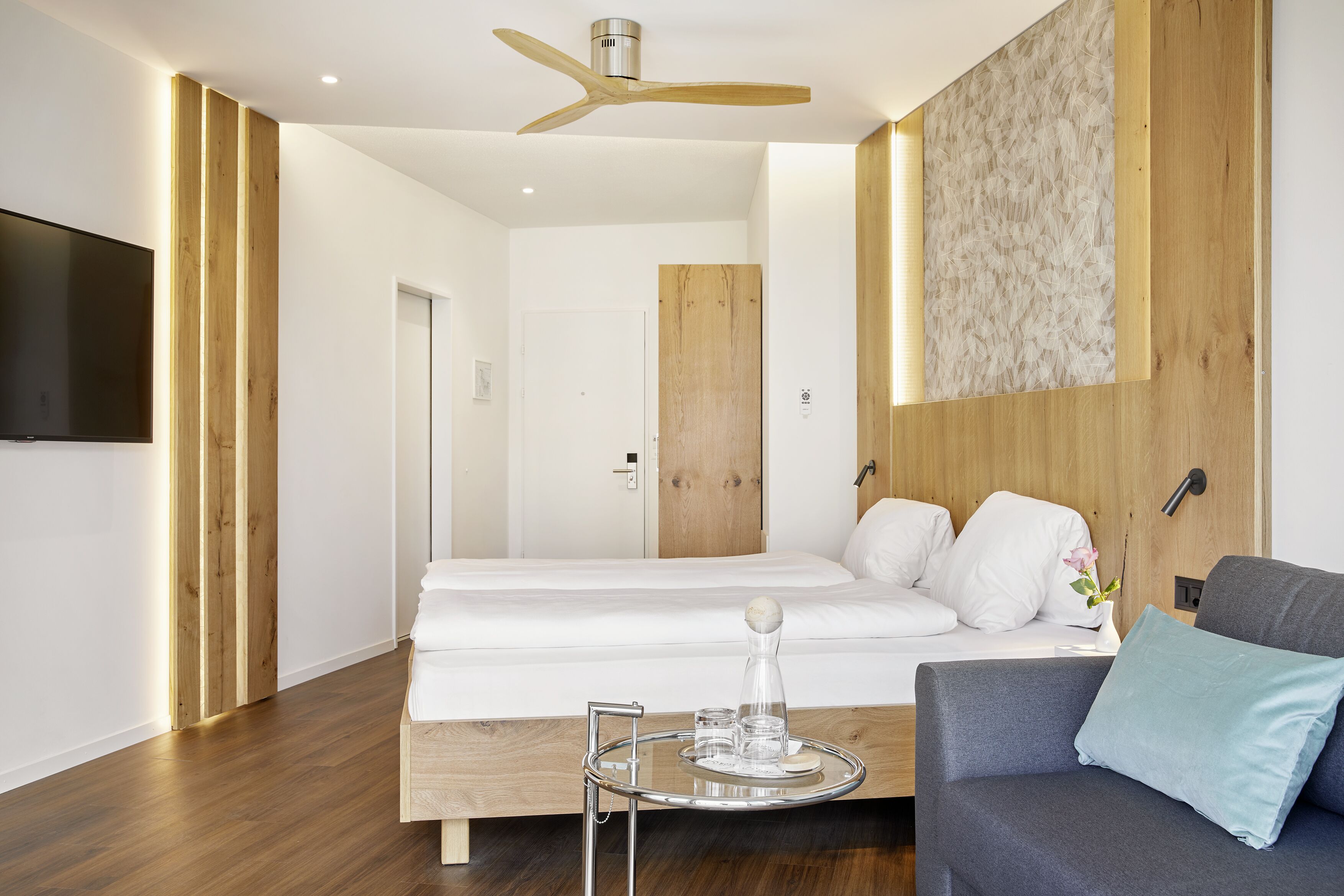 Ein modernes Hotelzimmer mit Deckenventilator aus Holz, einem großen Bett mit weißer Bettwäsche, einem grauen Sofa und einem kleinen runden Tisch mit Glasplatte.