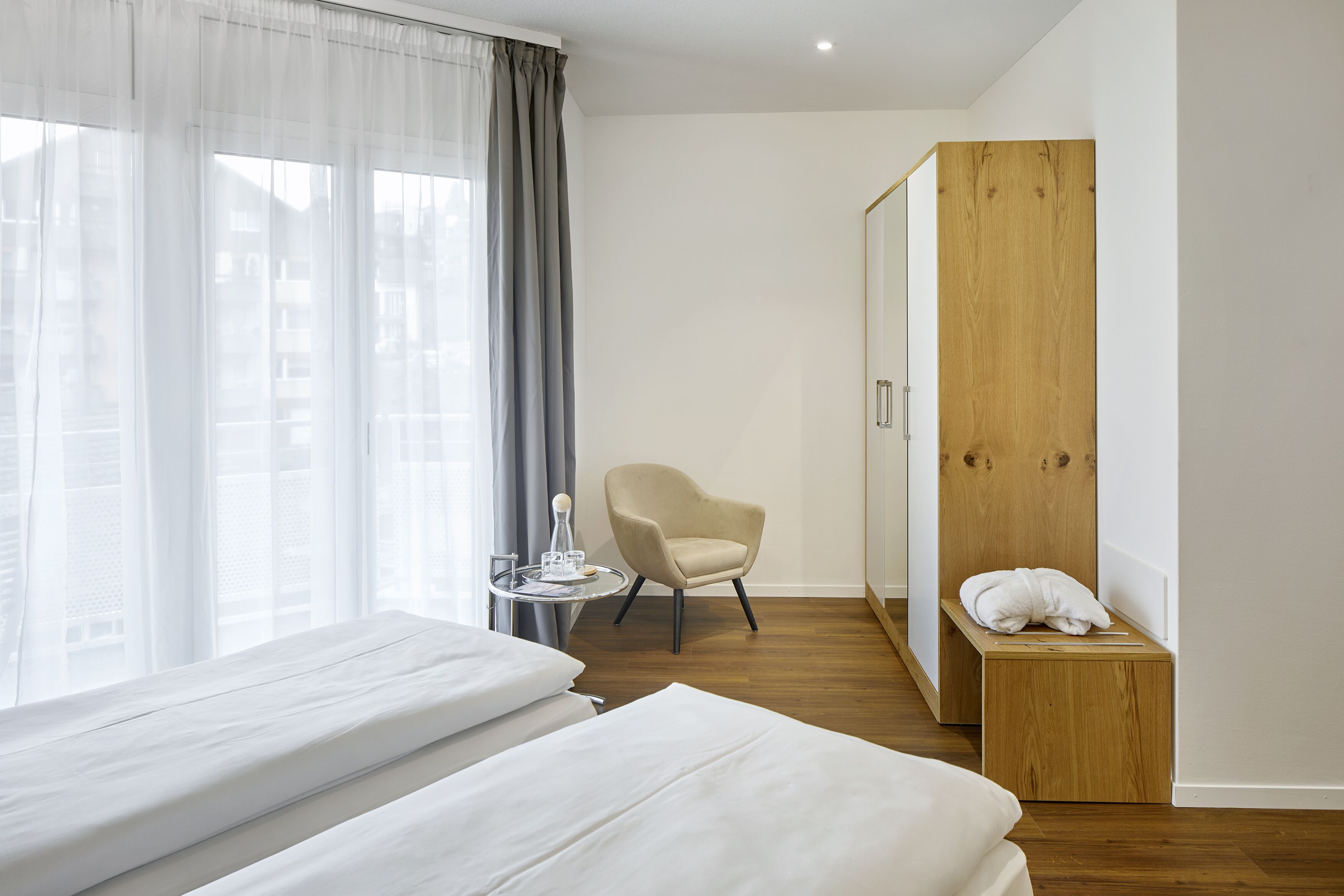 Ein Hotelzimmer mit einem Bett, einem Sessel, einem kleinen Tisch mit Glas und Flasche sowie einer Holzbank mit Handtuch.