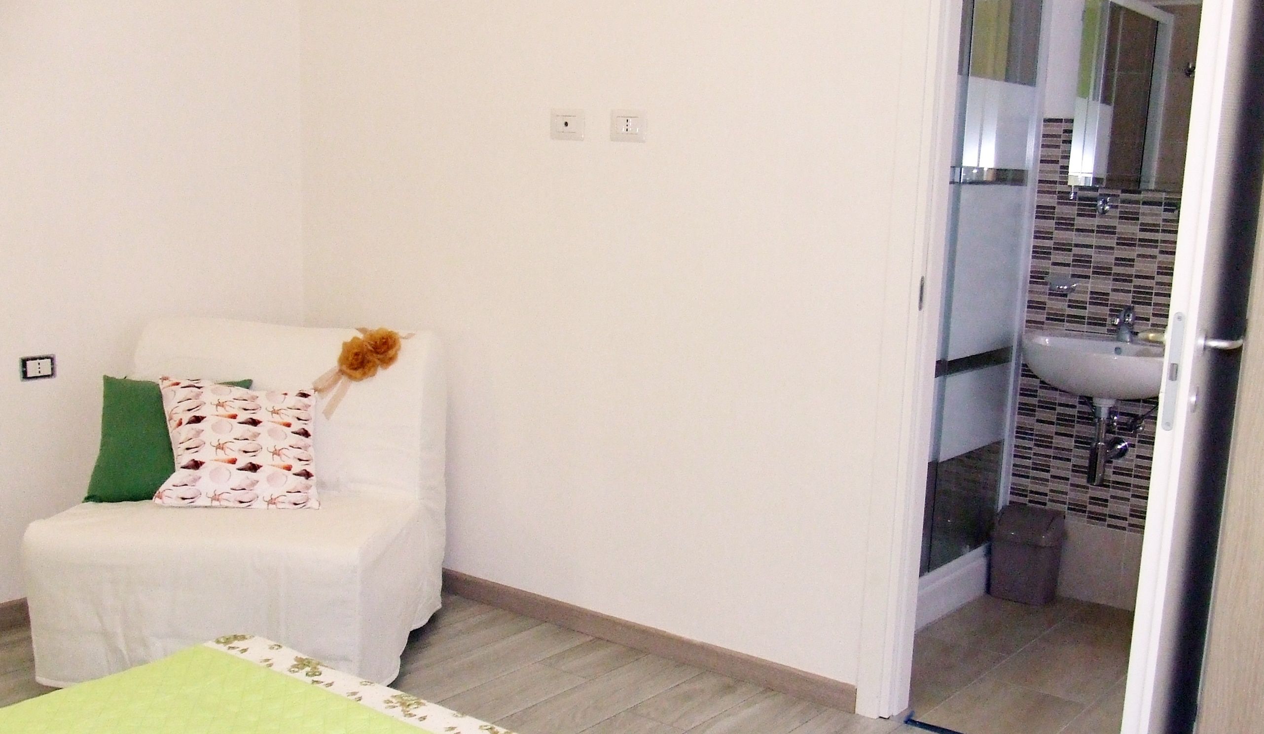 A small, clean room with a single bed, a green pillow, and a white chair with a floral cushion. A bathroom with a sink and tiled walls is visible through an open door.