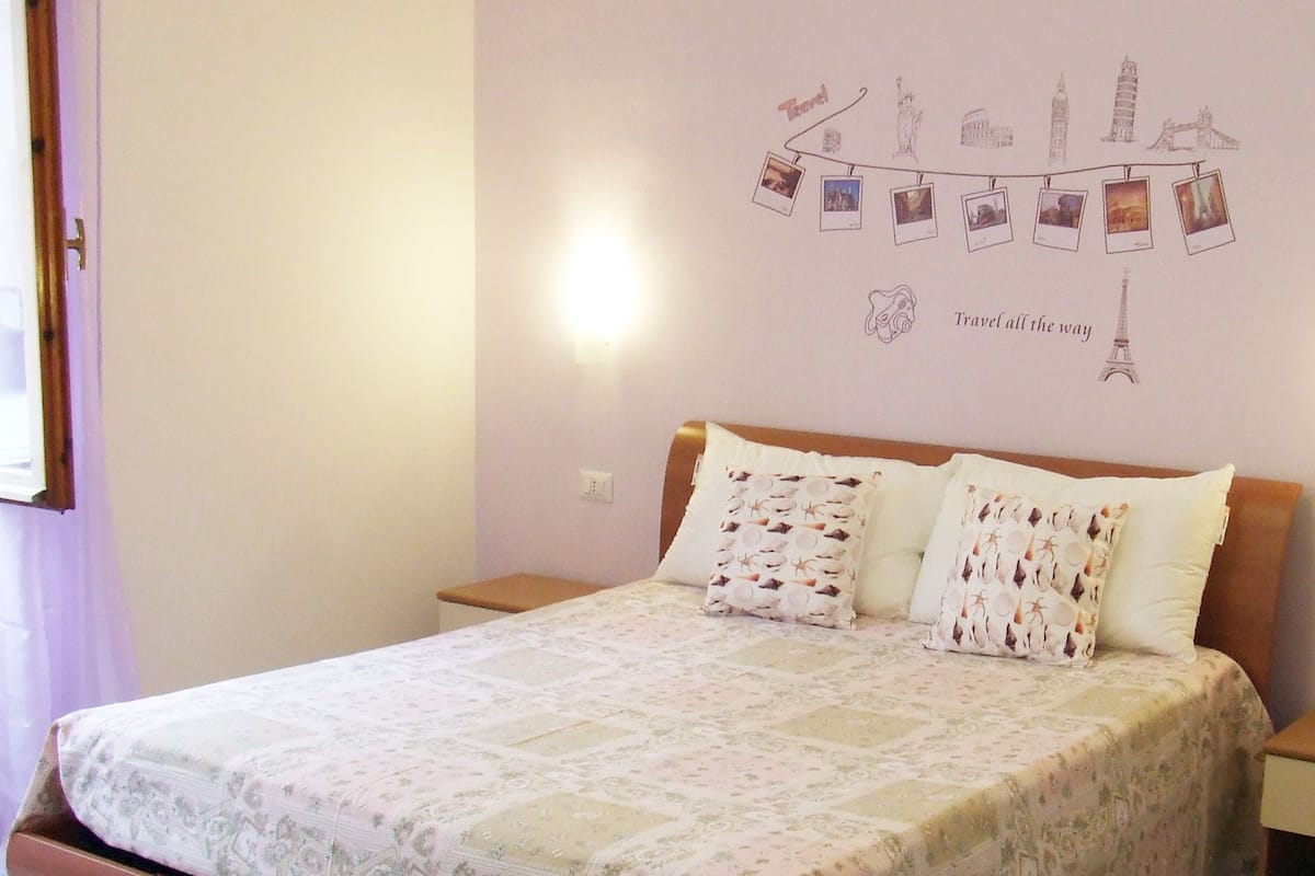 A bedroom with a bed, two pillows, a nightstand, a wall with a travel-themed decoration, and a window with curtains.