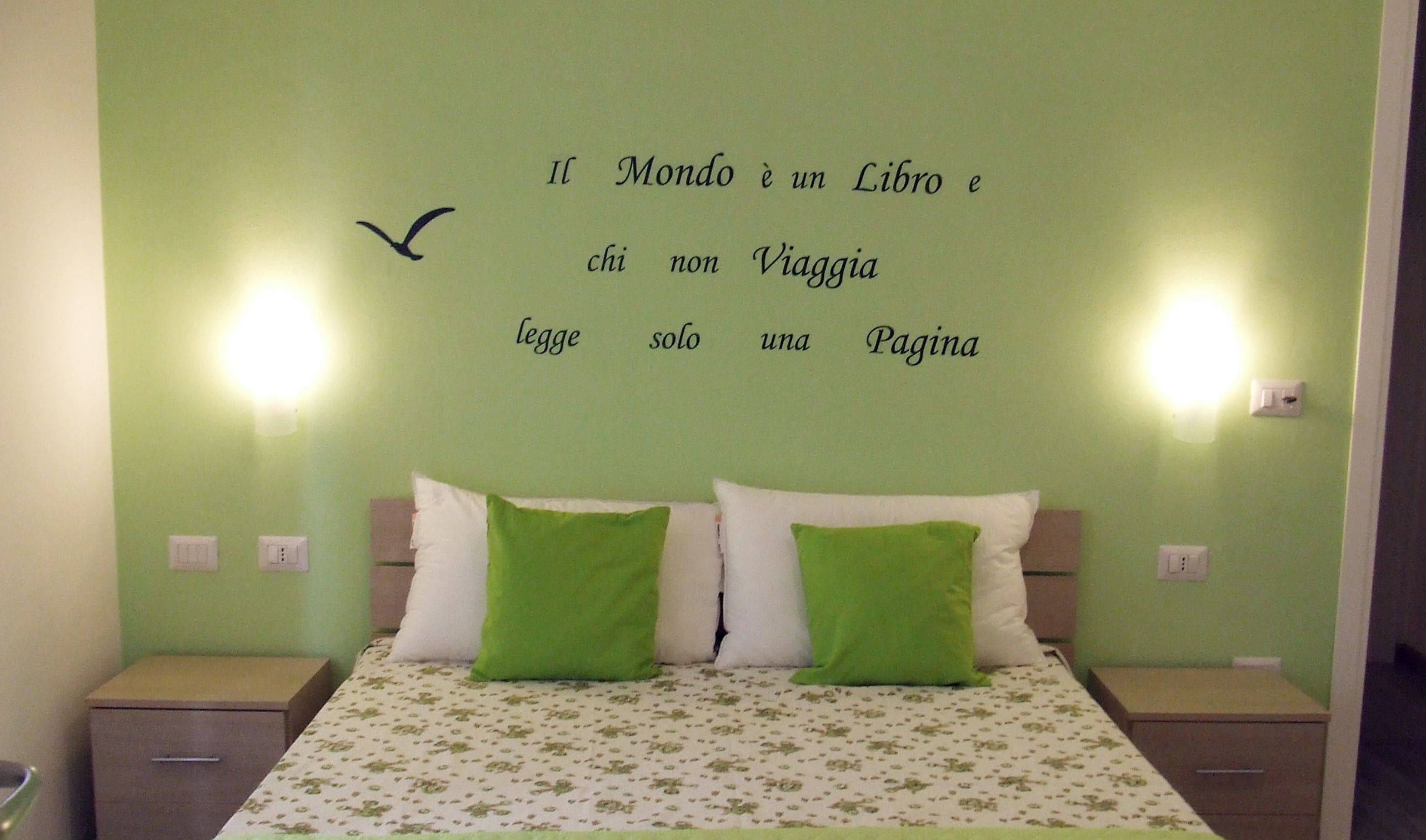 Une chambre à coucher avec un lit, deux oreillers verts, un mur orné d’une citation en italien et deux lampes fixées au mur.