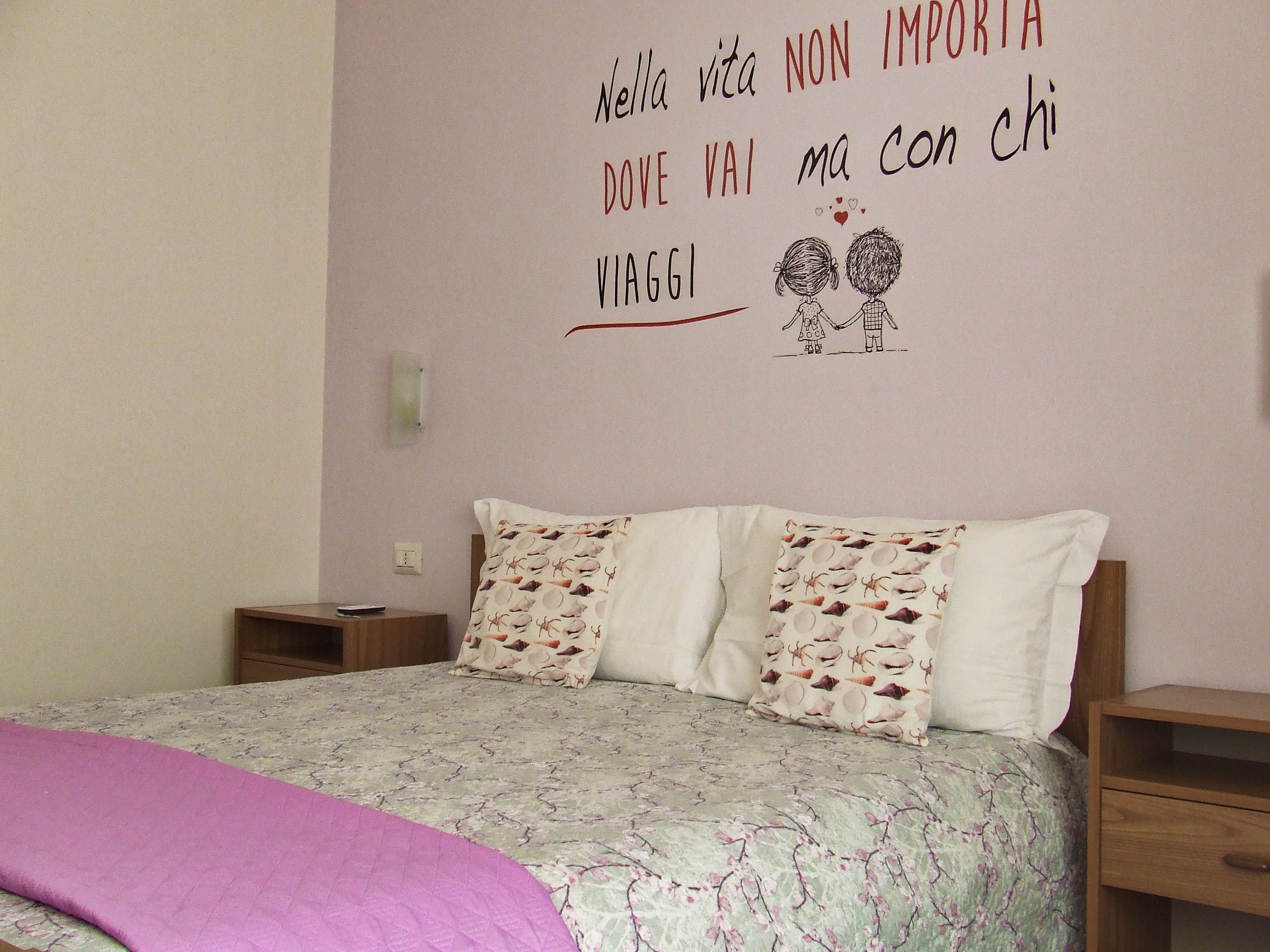A bedroom with a bed, two pillows, a bedside table, and a wall with a motivational quote and a drawing of two children holding hands.