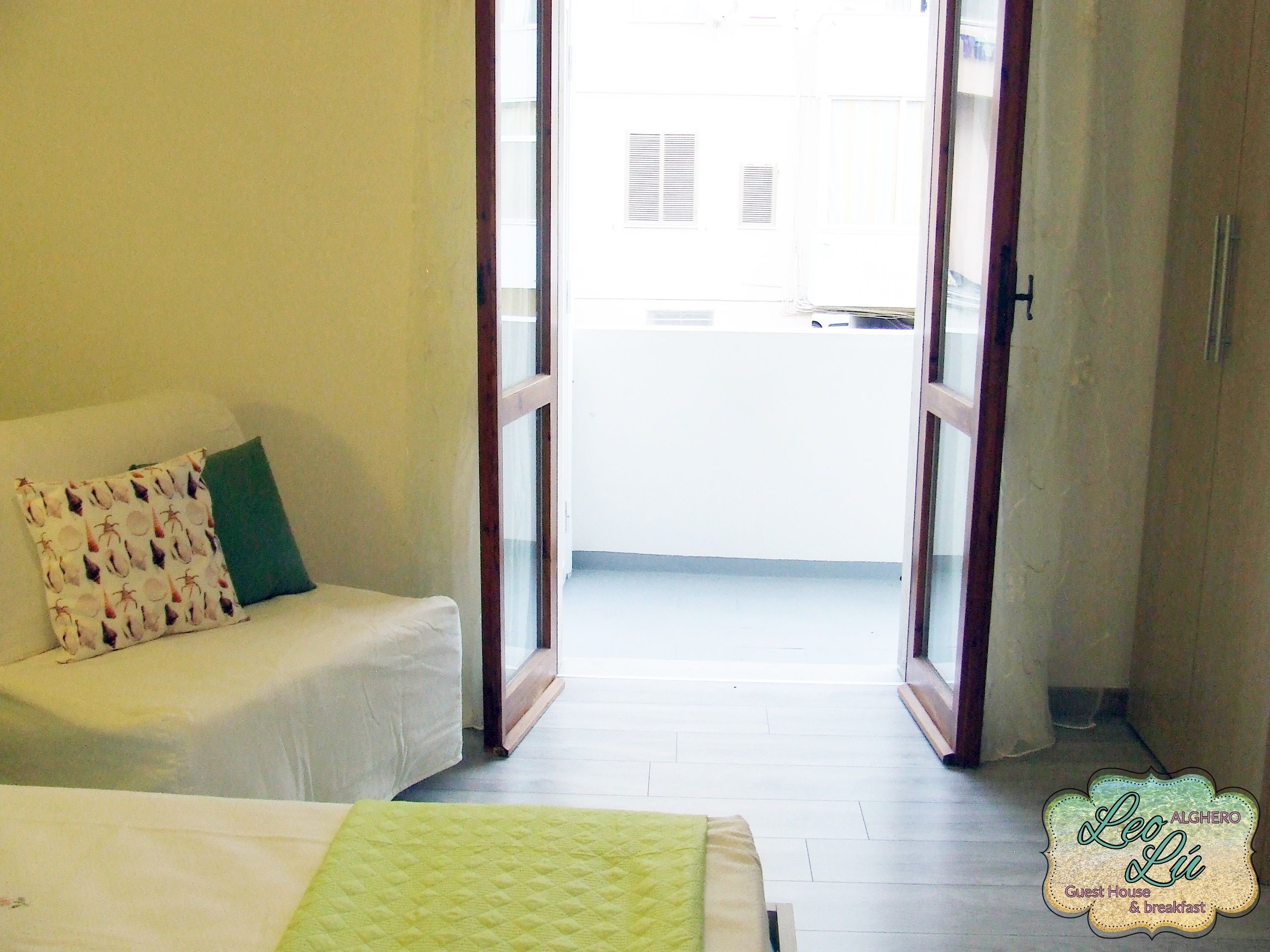 A room with a bed, a small sofa with patterned cushions, and a window with a view of a building.