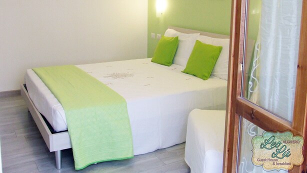 A single-bed room with a green bedspread, white bedding, and two green pillows. A wall-mounted light is visible, and a painting of a sunset over the ocean hangs above the bed. - Leolù Guesthouse and breakfast (Alghero)