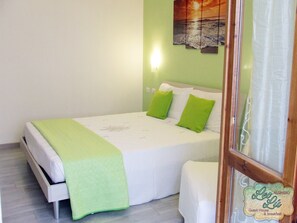A single-bed room with a green bedspread, white bedding, and two green pillows. A wall-mounted light is visible, and a painting of a sunset over the ocean hangs above the bed. - Leolù Guesthouse and breakfast (Alghero)
