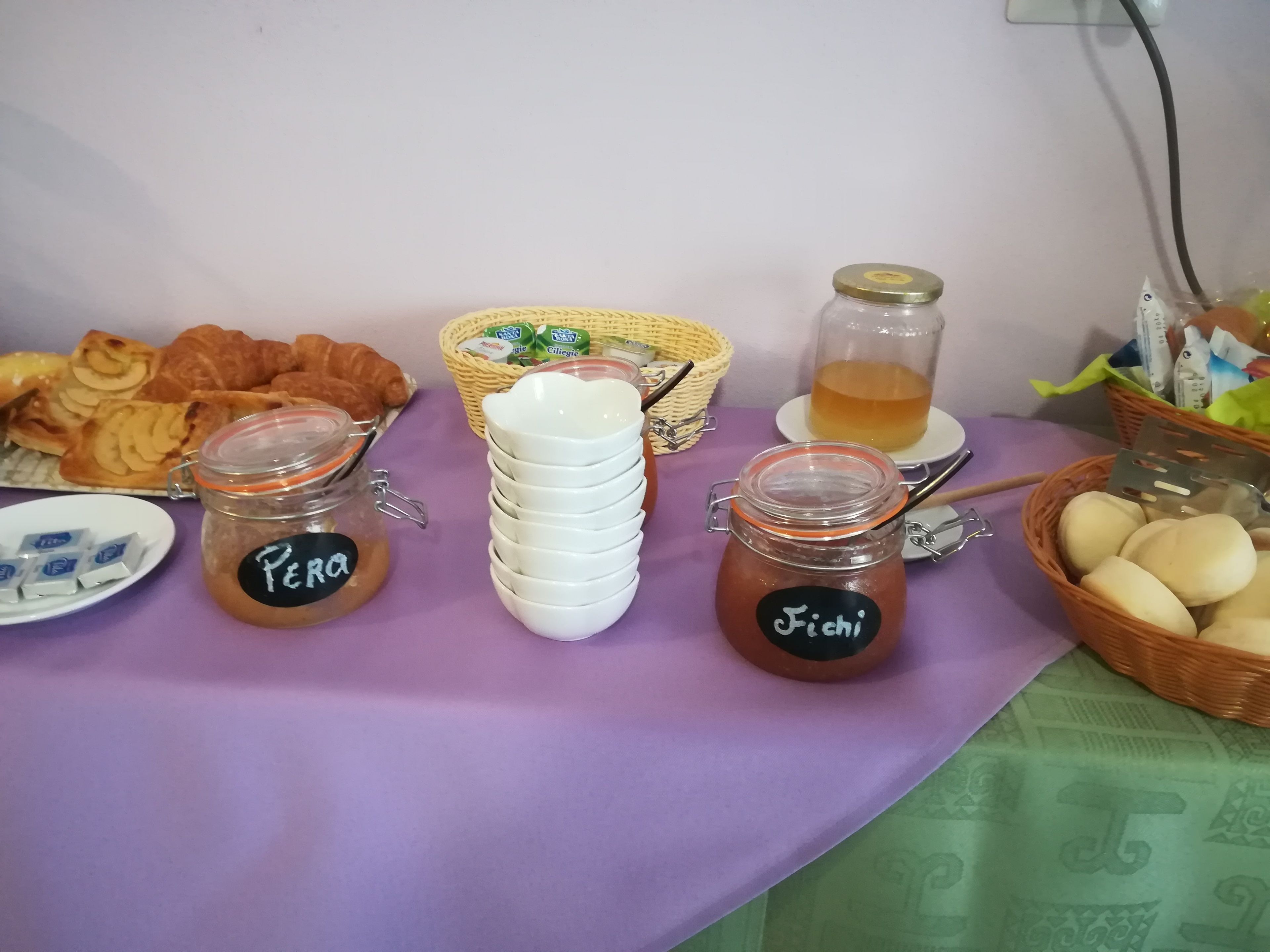 A table with food items including jars labeled "Perro" and "Fichi", a basket of bread, and a glass of tea.