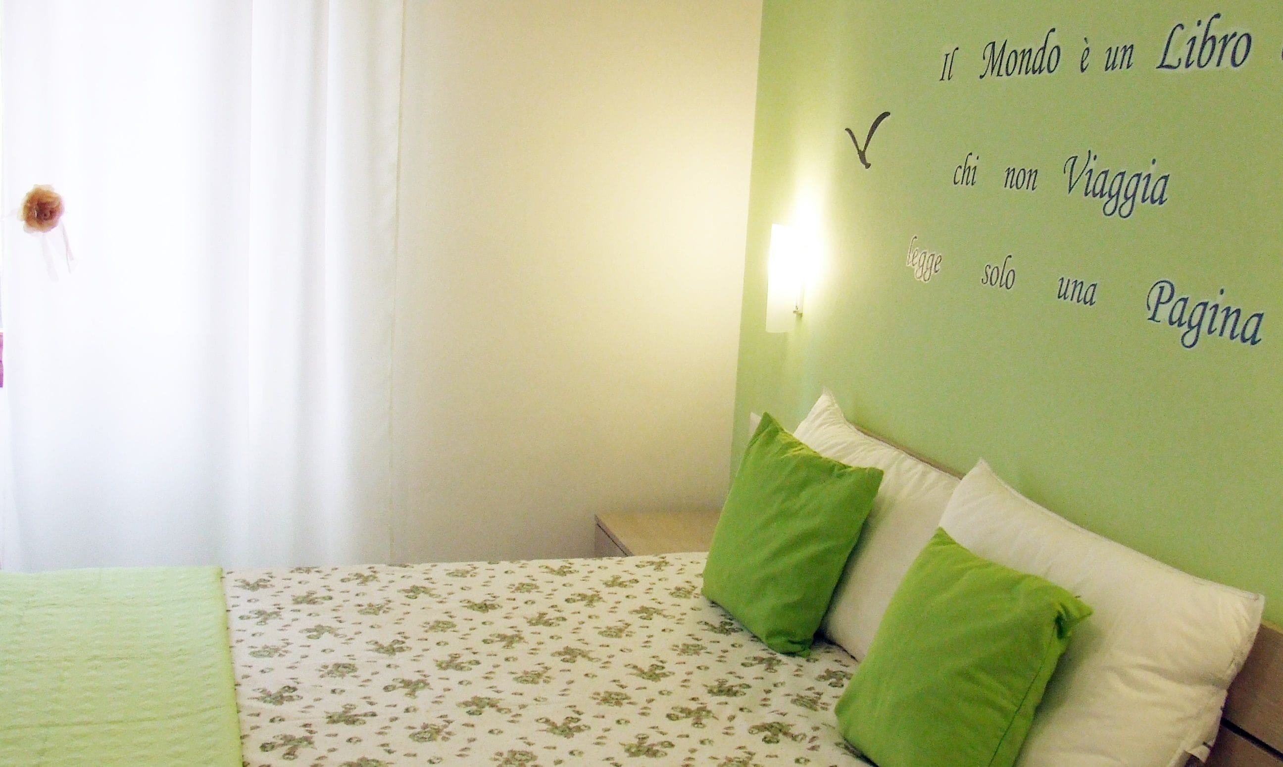 Quarto com cama, almofadas verdes e brancas, e parede com luminária e texto em italiano.
