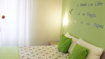 Quarto com cama, almofadas verdes e brancas, e parede com luminária e texto em italiano.