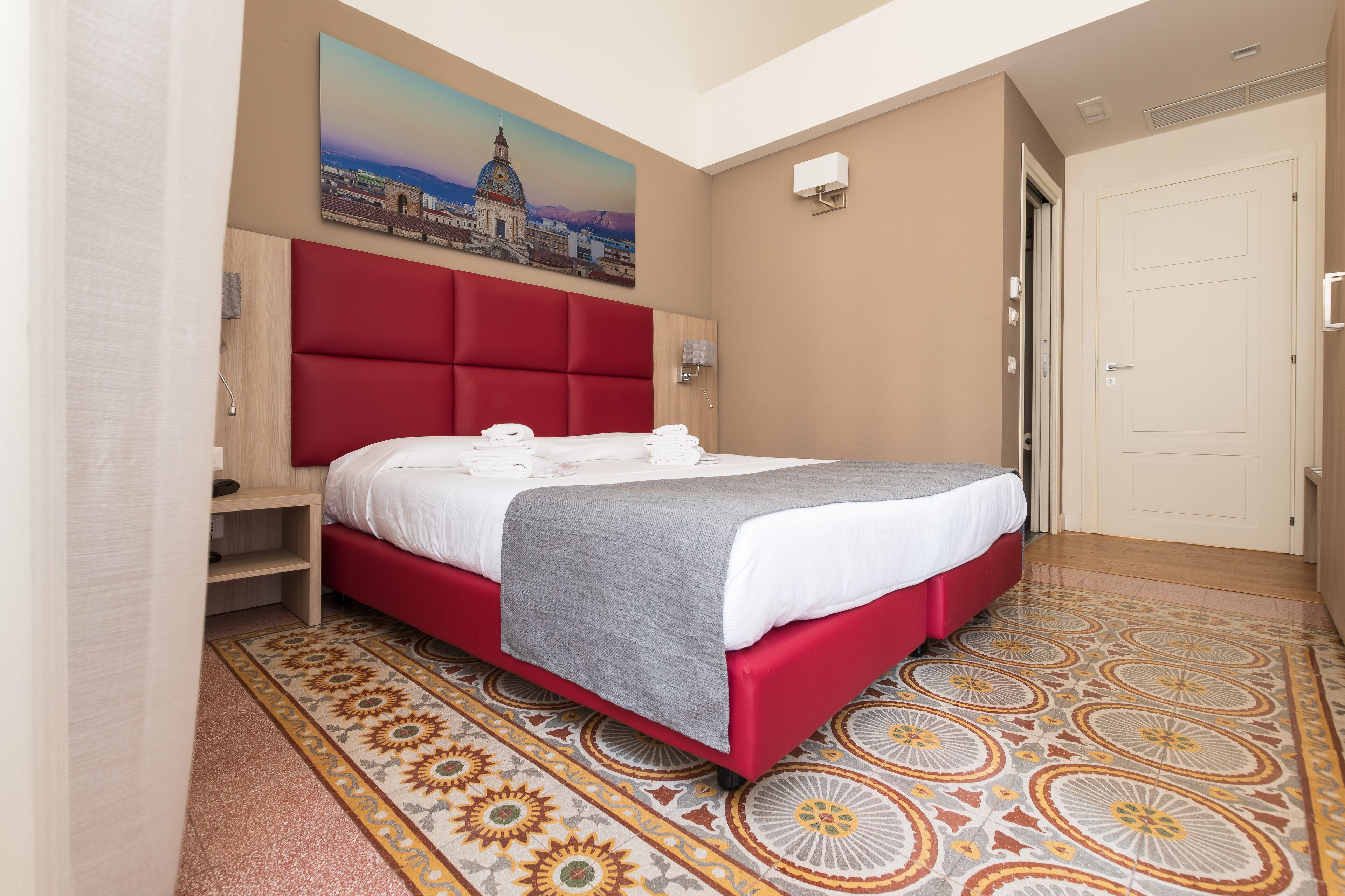 A hotel room with a large bed, a red headboard, a painting of a cityscape, a patterned floor, and a door.