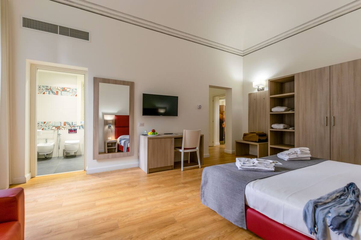 Ett modernt hotellrum med trägolv, en säng med ett grått överkast, ett skrivbord med en stol, en tv som är monterad på väggen och en garderob.