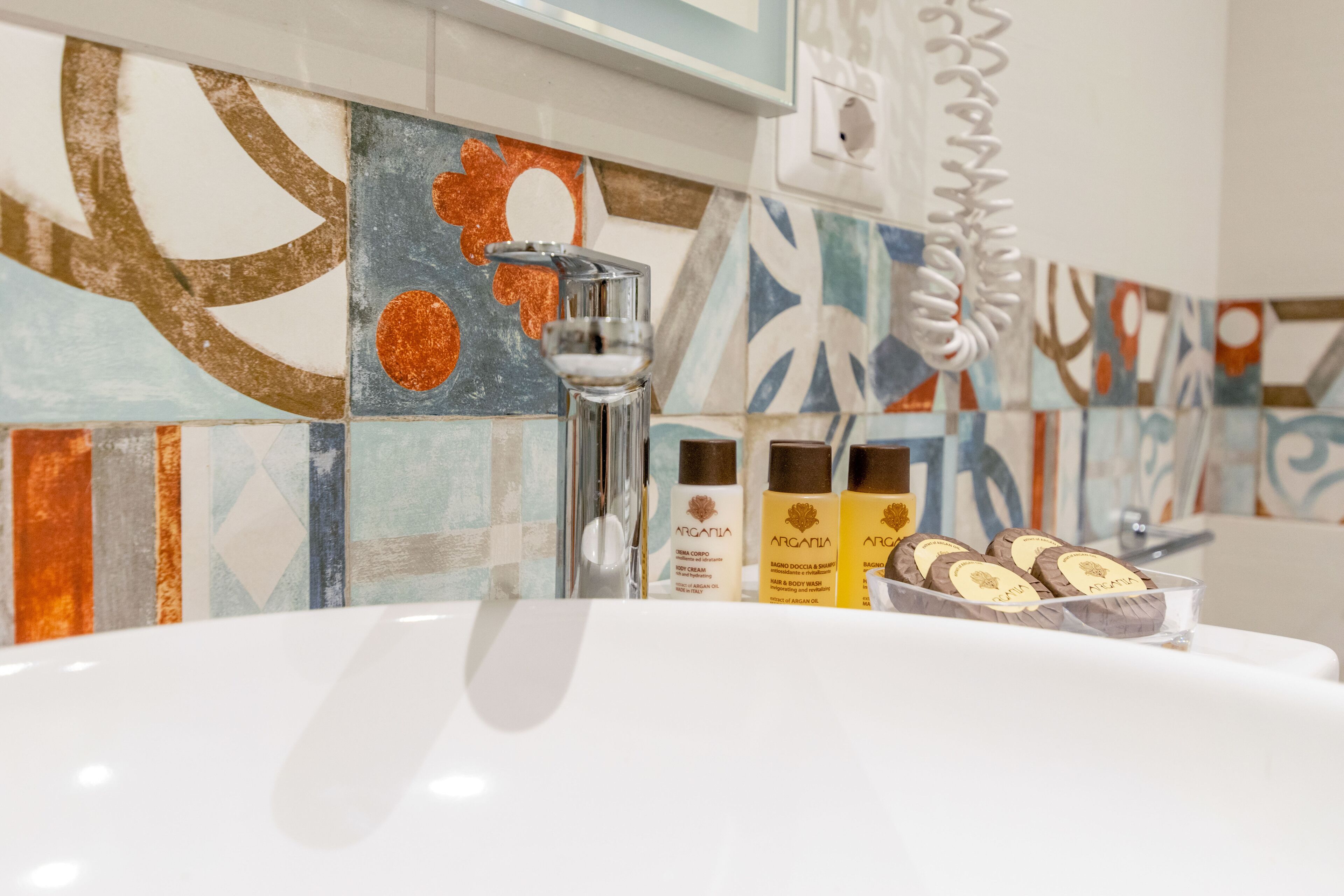 Un baño con un lavamanos blanco, baldosas decorativas y tres botellas de jabón corporal Argania.