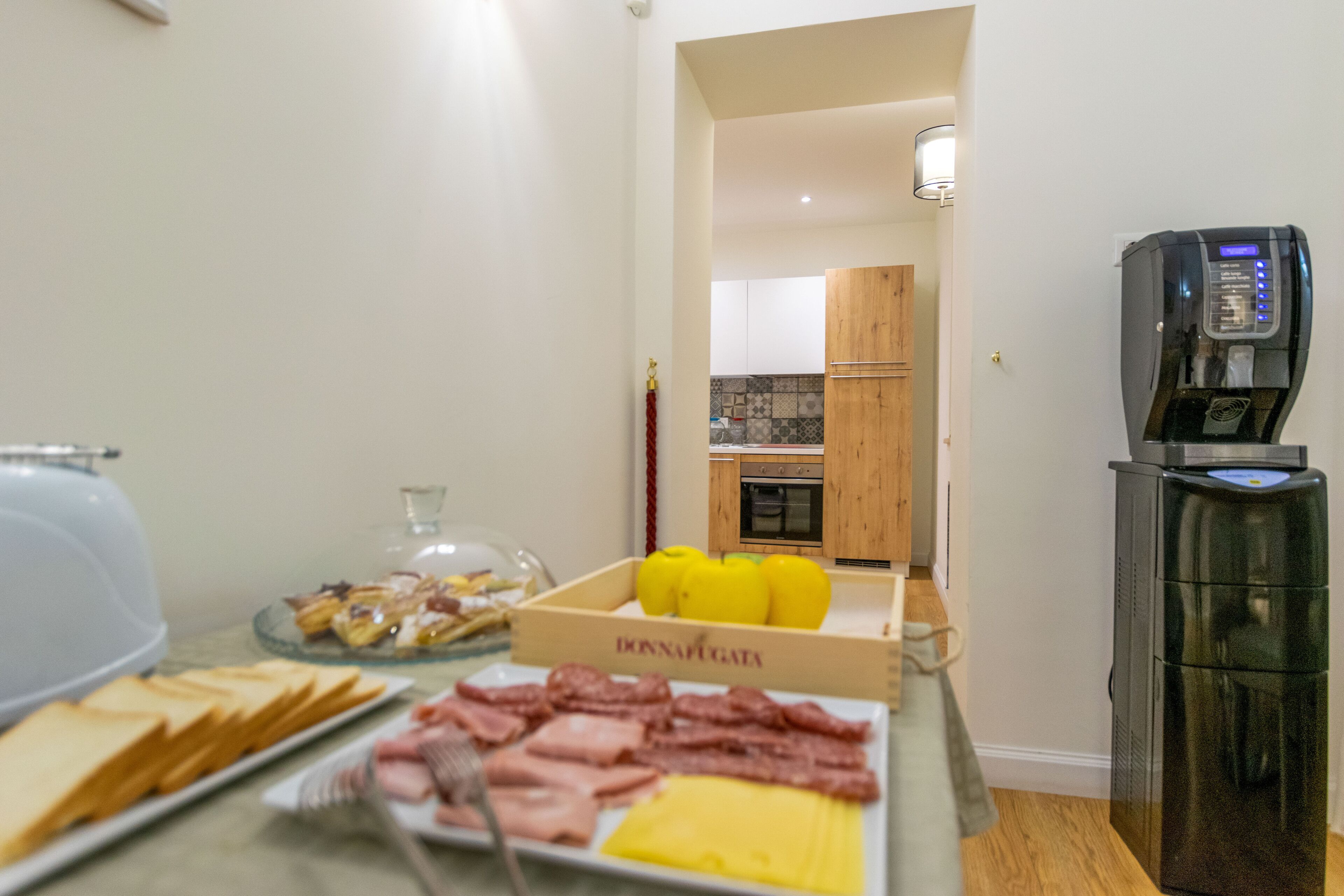 A breakfast buffet with various food items, a coffee machine, and a kitchen area in the background.