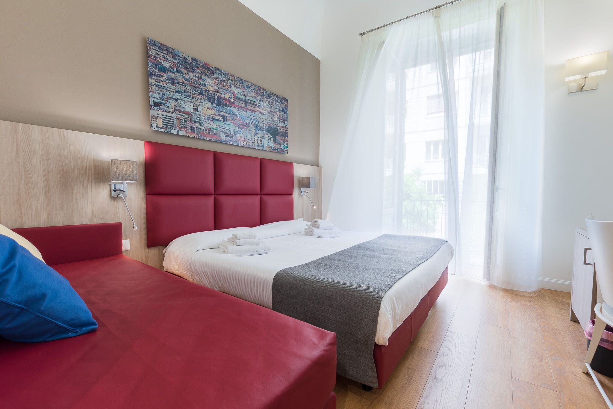 A hotel room with two beds, a red headboard, a cityscape painting, a window with sheer curtains, and a bedside lamp.