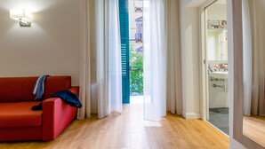 Una habitación con sofá rojo, suelo de madera, baño con lavabo y vista a un balcón a través de una puerta.