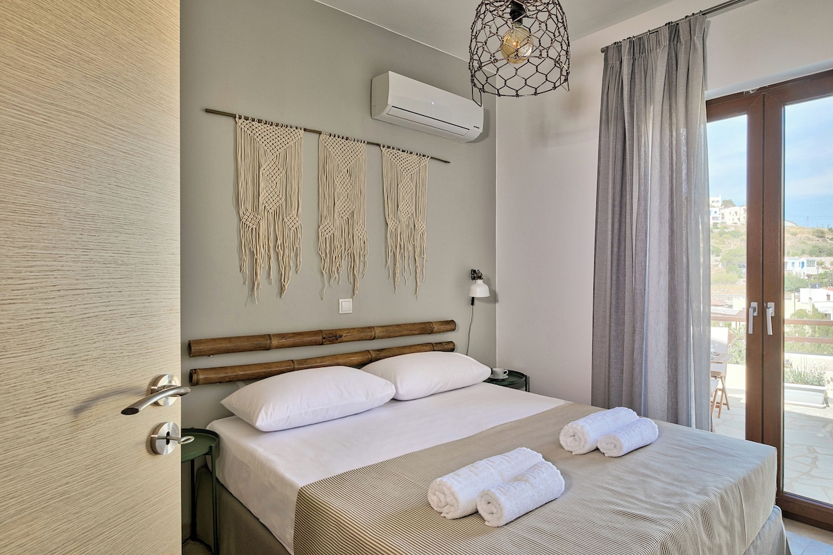 A hotel room with a bed, macrame wall hanging, an air conditioner, a nightstand, and a view of the outside.