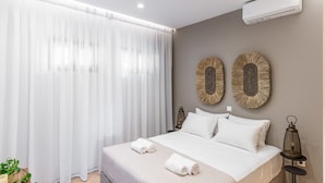 A modern bedroom with a large bed, white linens, a bedside table with a lantern, and two wall-mounted decorative items. - Syra Suites (Syros)