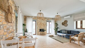 A spacious living area with a stone wall, a stone fireplace, a sofa, and a dining table with chairs. - Syra Suites (Syros)
