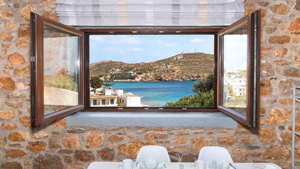 A dining area with a table set for two, a stone wall, and a window with an open shutter revealing a coastal view. - Syra Suites (Syros)