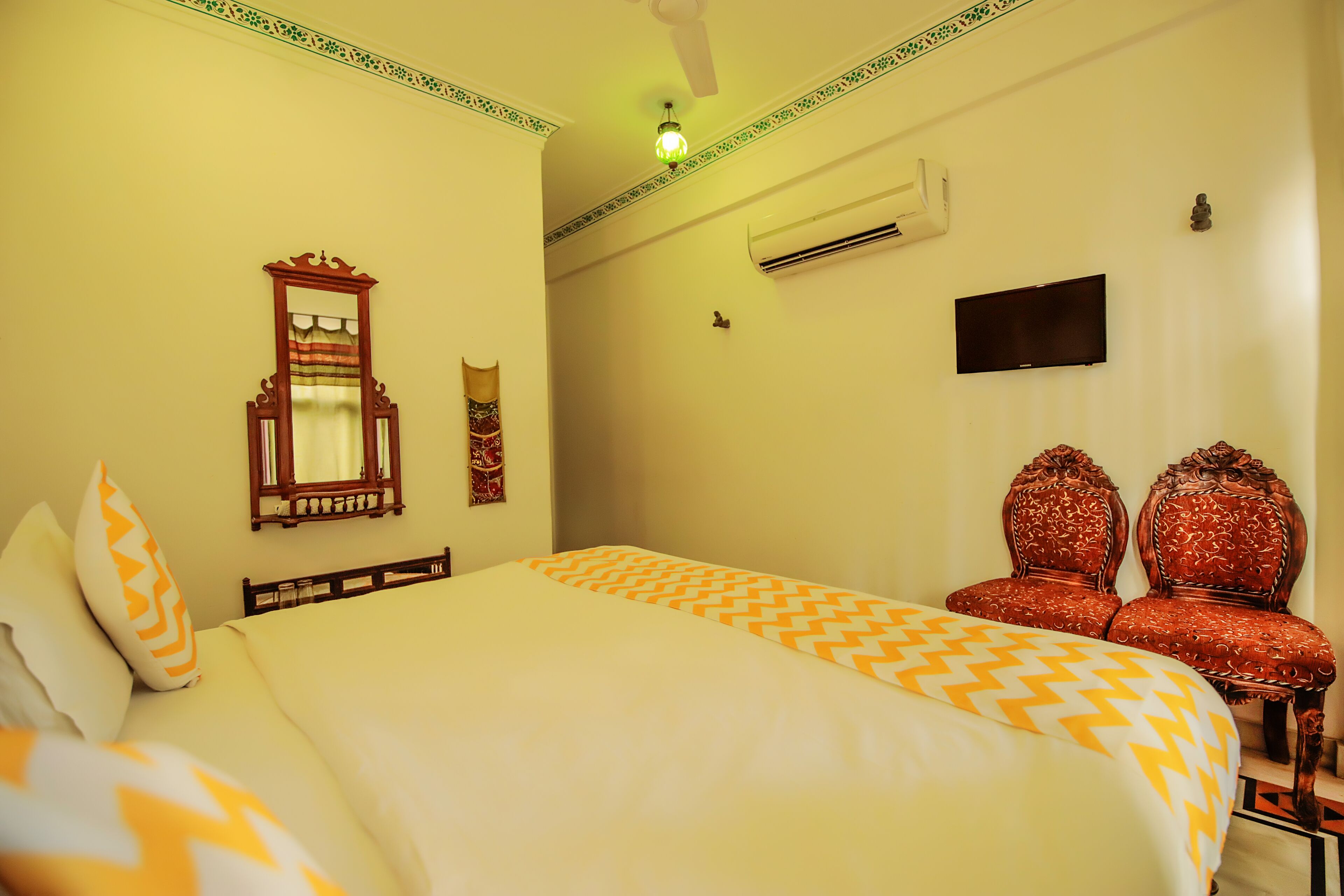 A hotel room with a bed, two ornate chairs, a mirror, an air conditioning unit, and a television.