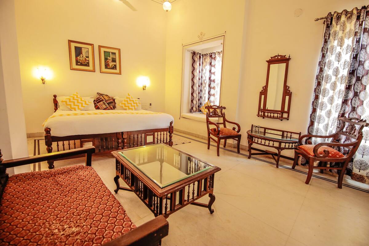 A hotel room with a bed, a glass-top table, two wooden chairs, a sofa, a mirror, and two wall-mounted lamps.
