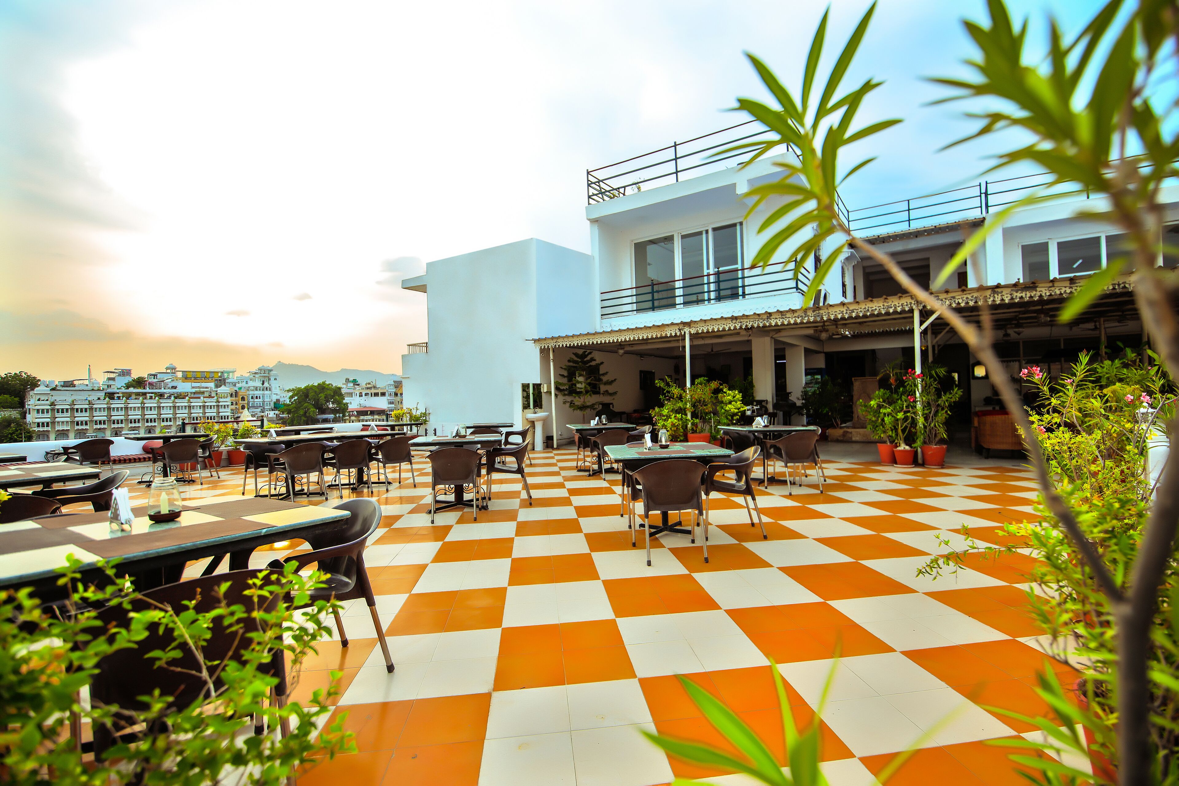 Un espace de restauration en plein air doté d’un sol à carreaux, de tables et de chaises, entouré de plantes en pot et offrant une vue sur le paysage urbain.