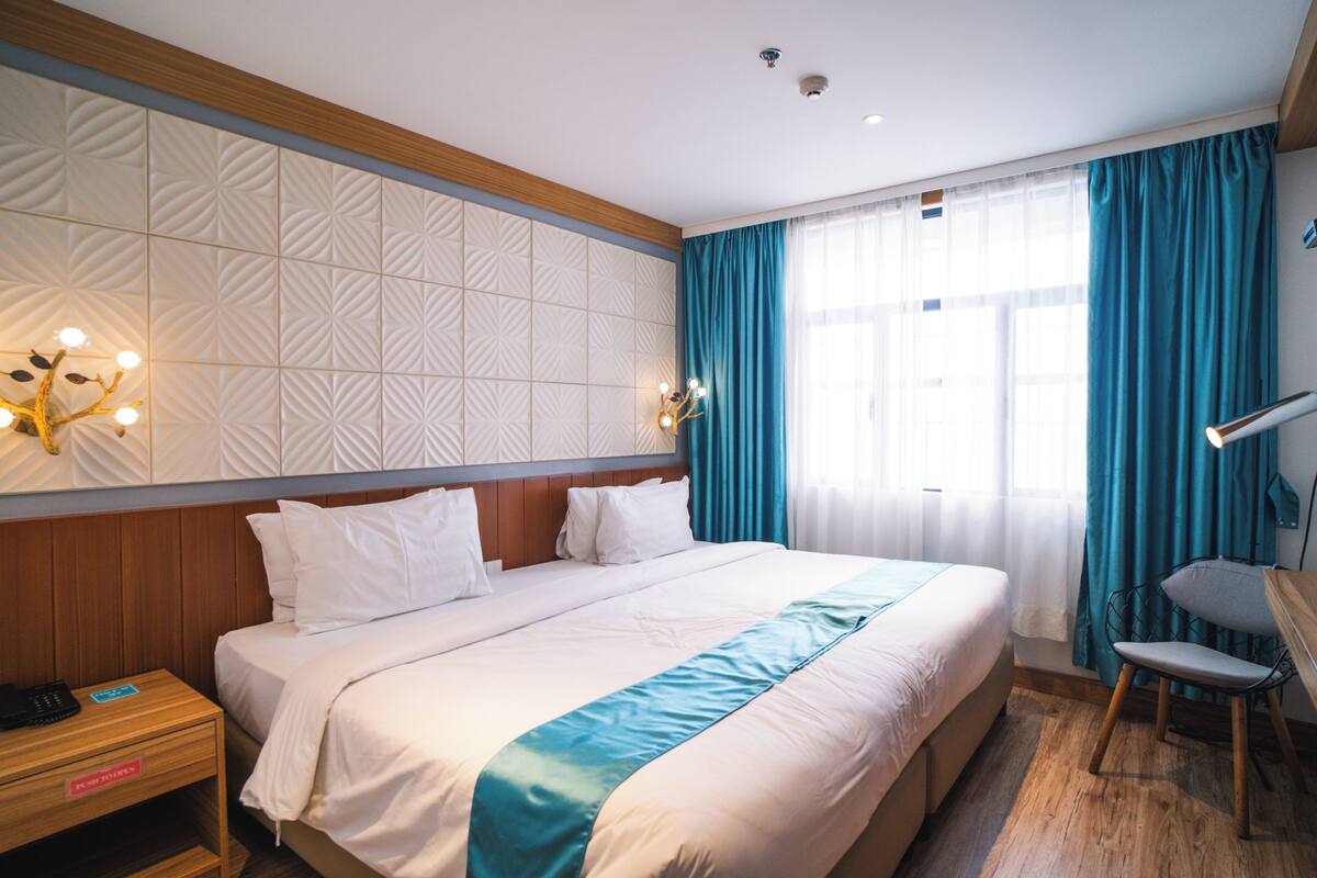 Une chambre d’hôtel avec un grand lit, une table de chevet, un bureau avec une chaise et une fenêtre avec des rideaux.