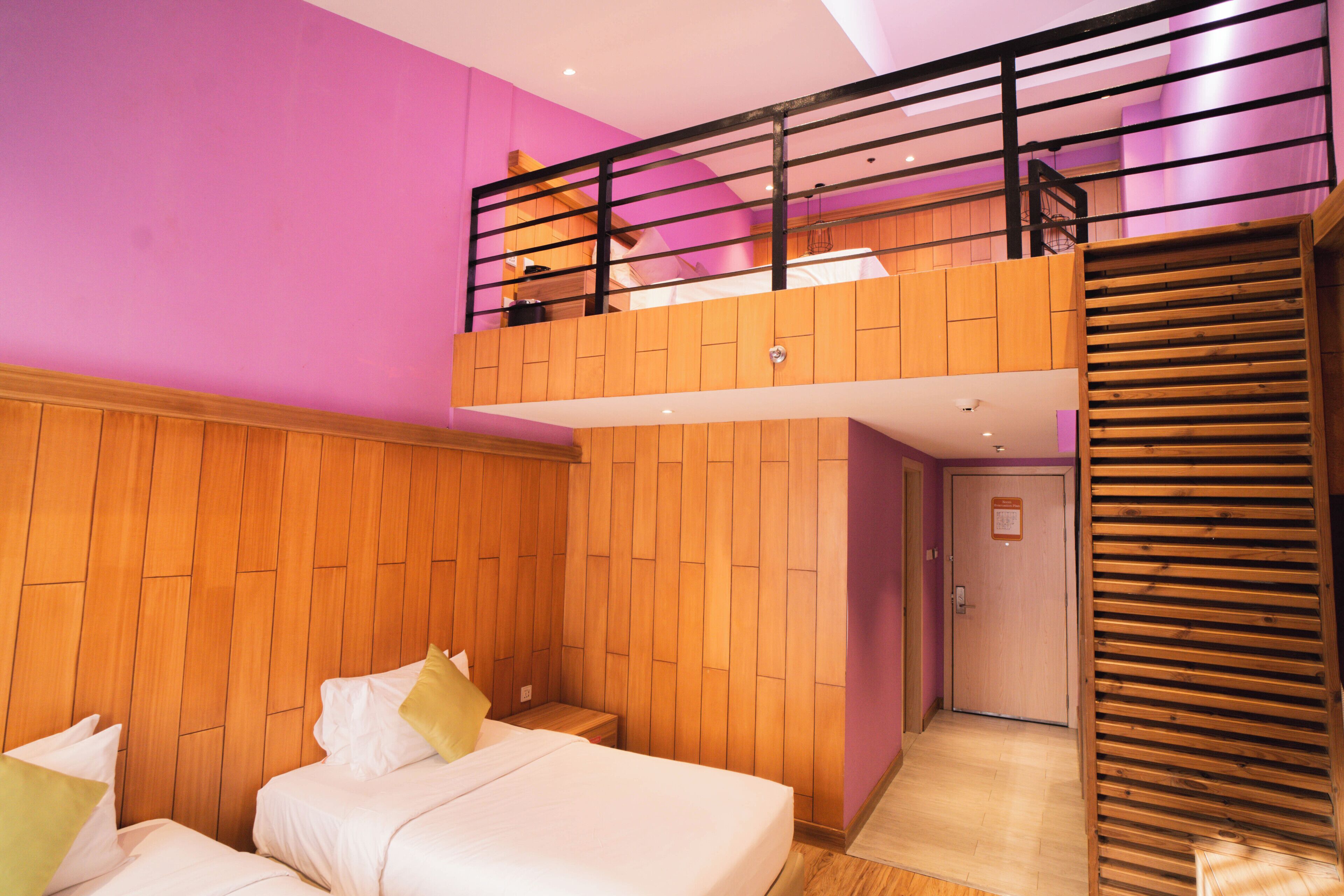 A hotel room with a wooden floor, a bed with pillows, a wooden wall panel, a balcony with a railing, and a door.