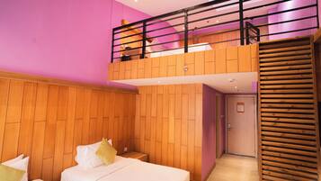 A hotel room with a wooden floor, a bed with pillows, a wooden wall panel, a balcony with a railing, and a door.