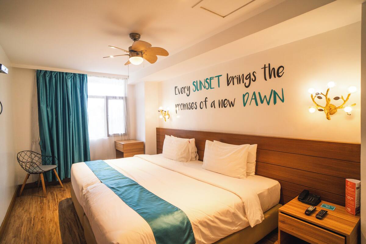 Une chambre d’hôtel comprenant un lit, une table de chevet avec un téléphone et une télécommande, une chaise, un ventilateur de plafond et un mur orné d’une citation inspirante.