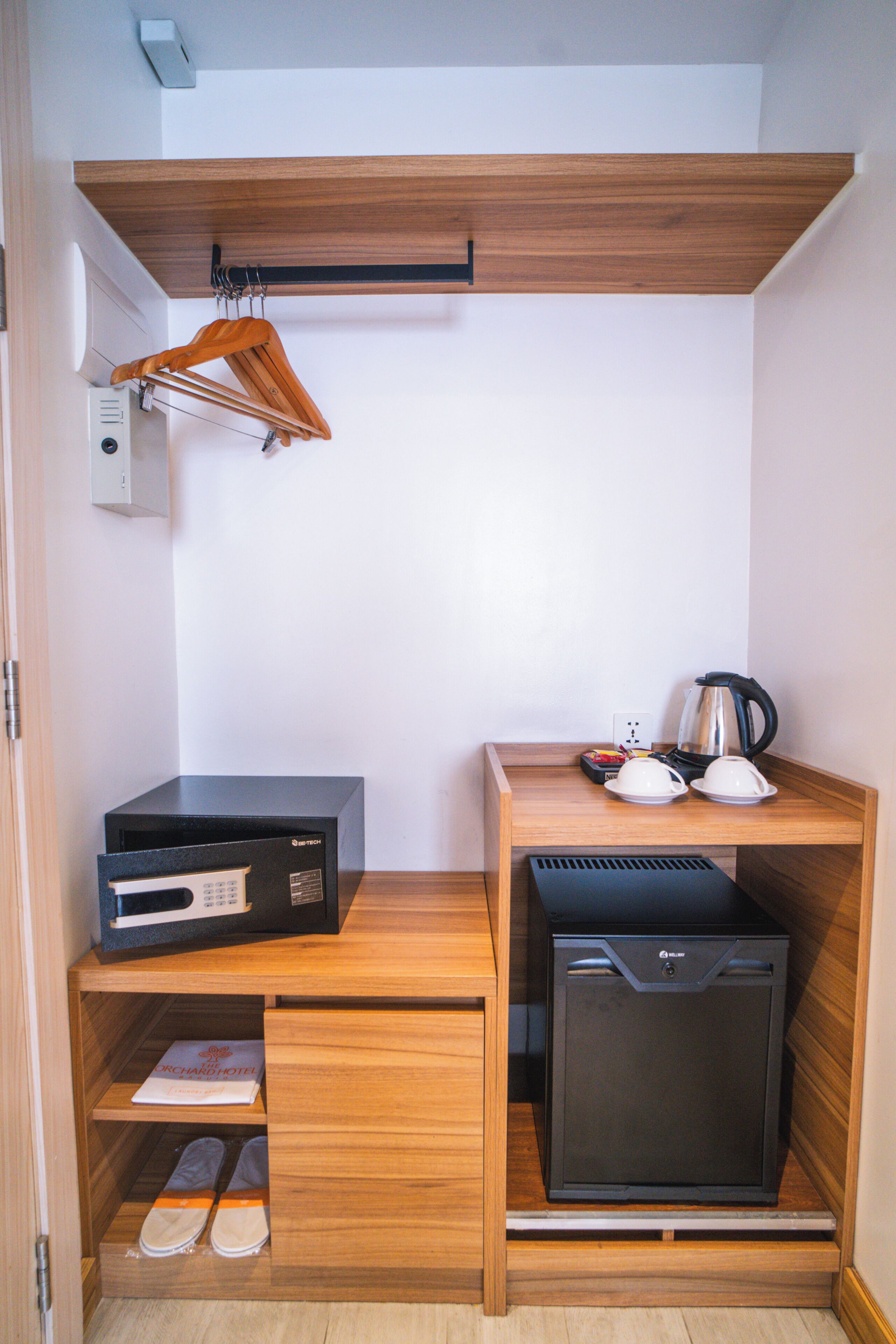 A compact kitchenette with a microwave, coffee maker, and a small refrigerator. There is a wooden shelf with a pair of shoes and a hanging garment rack.
