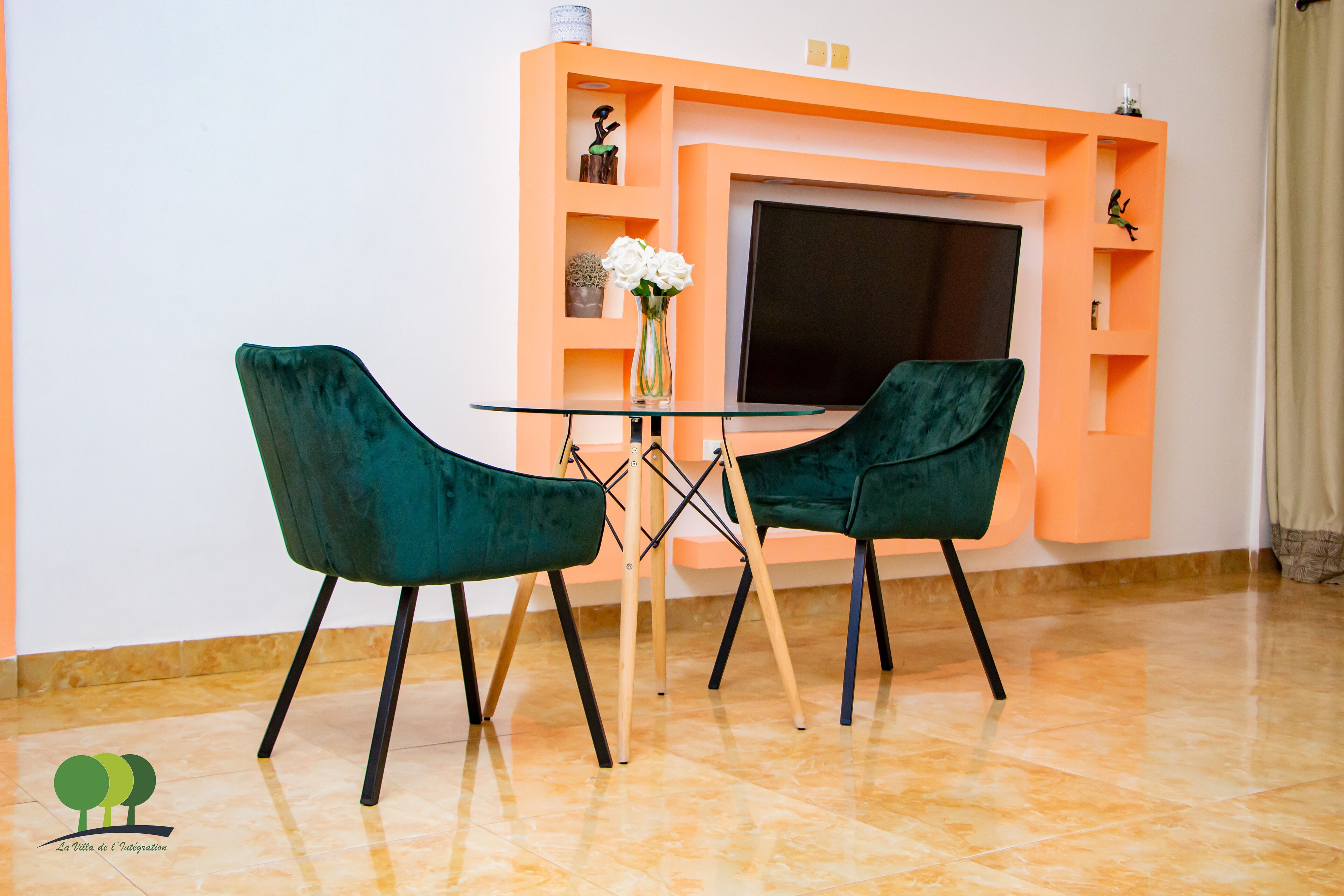 A room with a glass table and two green chairs, a TV, and a shelf with decorative items.