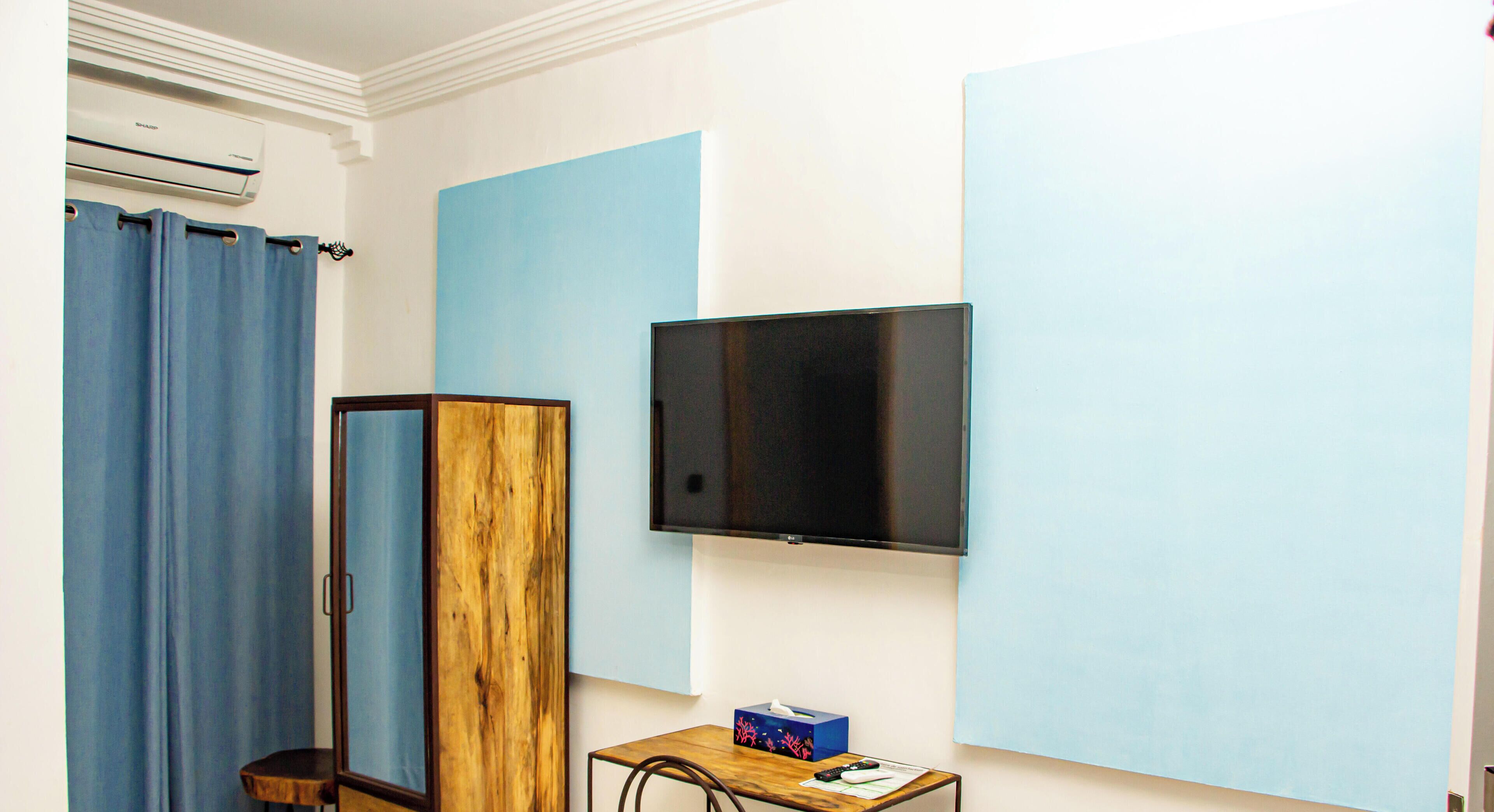 A room with a flat-screen TV, a wooden wardrobe, a small table with a box, and a blue curtain.