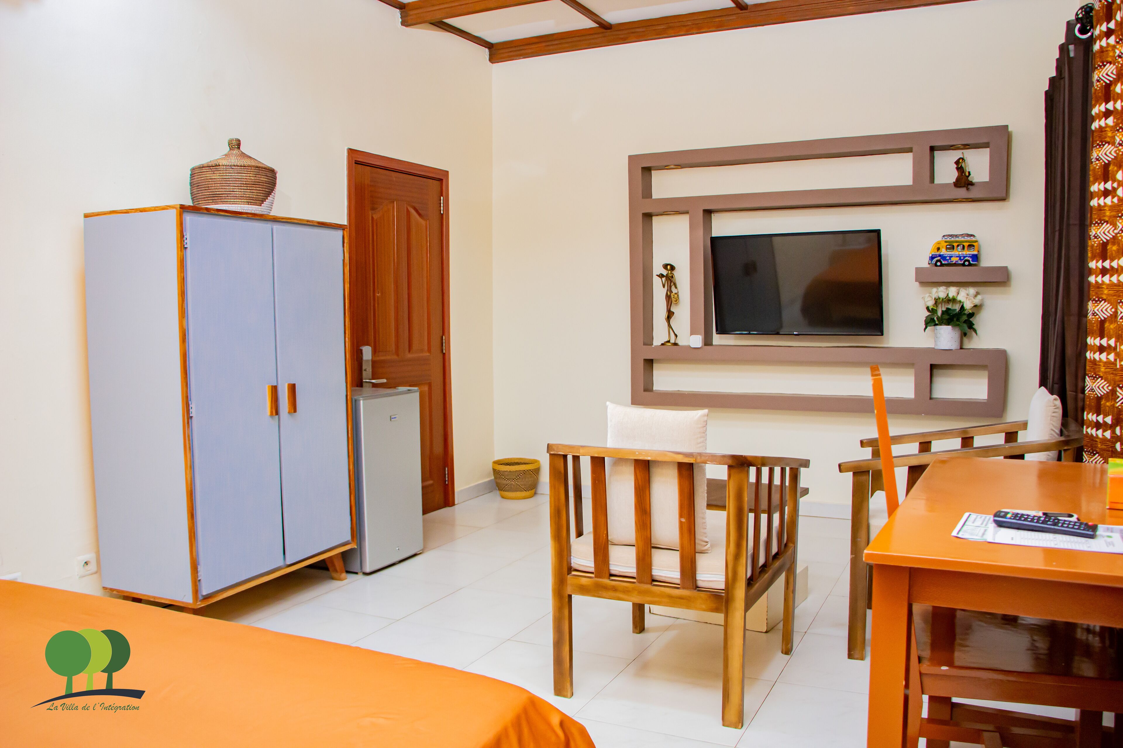 A room with a bed, a wooden chair, a table with a remote control, a television mounted on the wall, and a wardrobe.