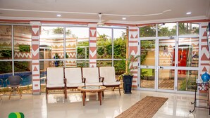 A spacious indoor area with a seating arrangement, a small table, and a large window with a view of greenery outside. - Villa De l'Intégration (Ouagadougou)