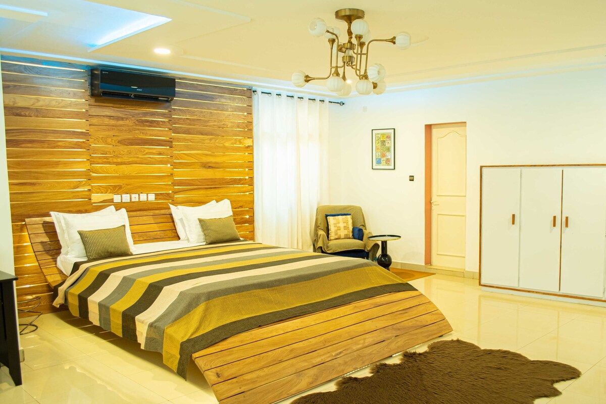 A modern bedroom with a wooden headboard, a large bed with striped bedding, a leather rug, a wardrobe, and a ceiling light fixture.