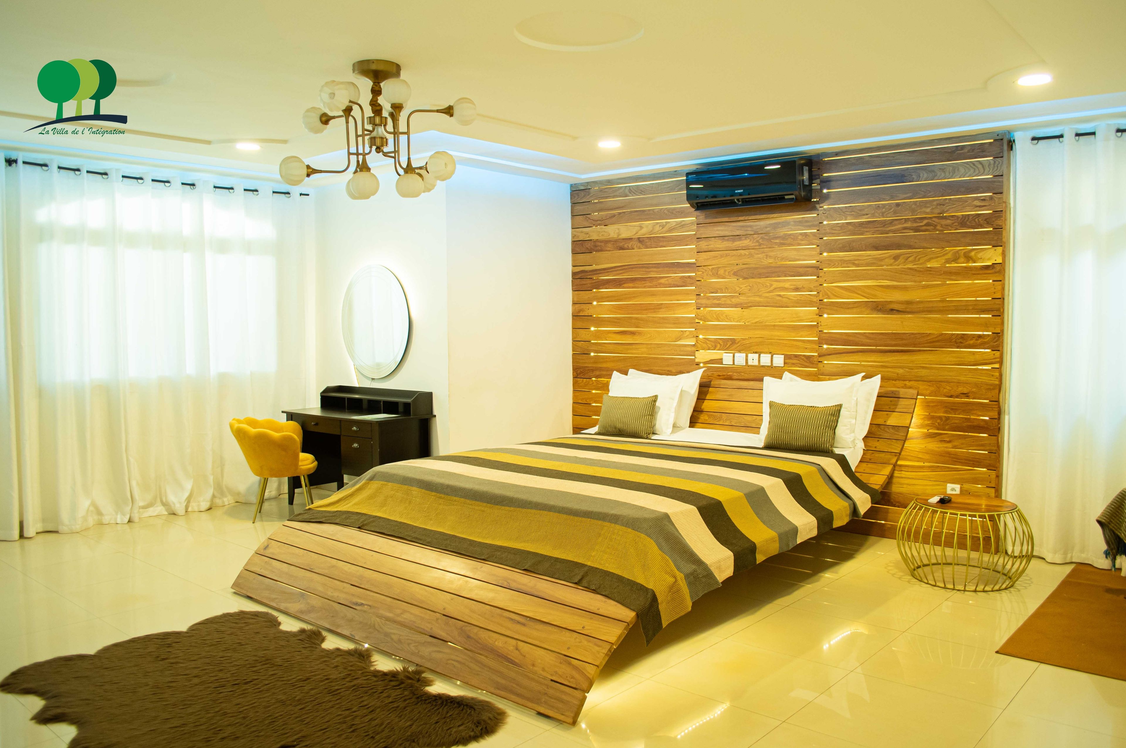 A modern bedroom with a wooden headboard, a striped bedspread, a round mirror, and a yellow chair.